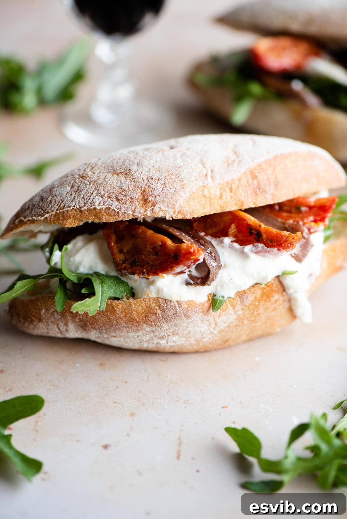 A side shot of a ciabatta sandwich with burrata, anchovies, sun blushed tomatoes and arugula on a light stone surface, emphasizing its freshness and artisanal quality.