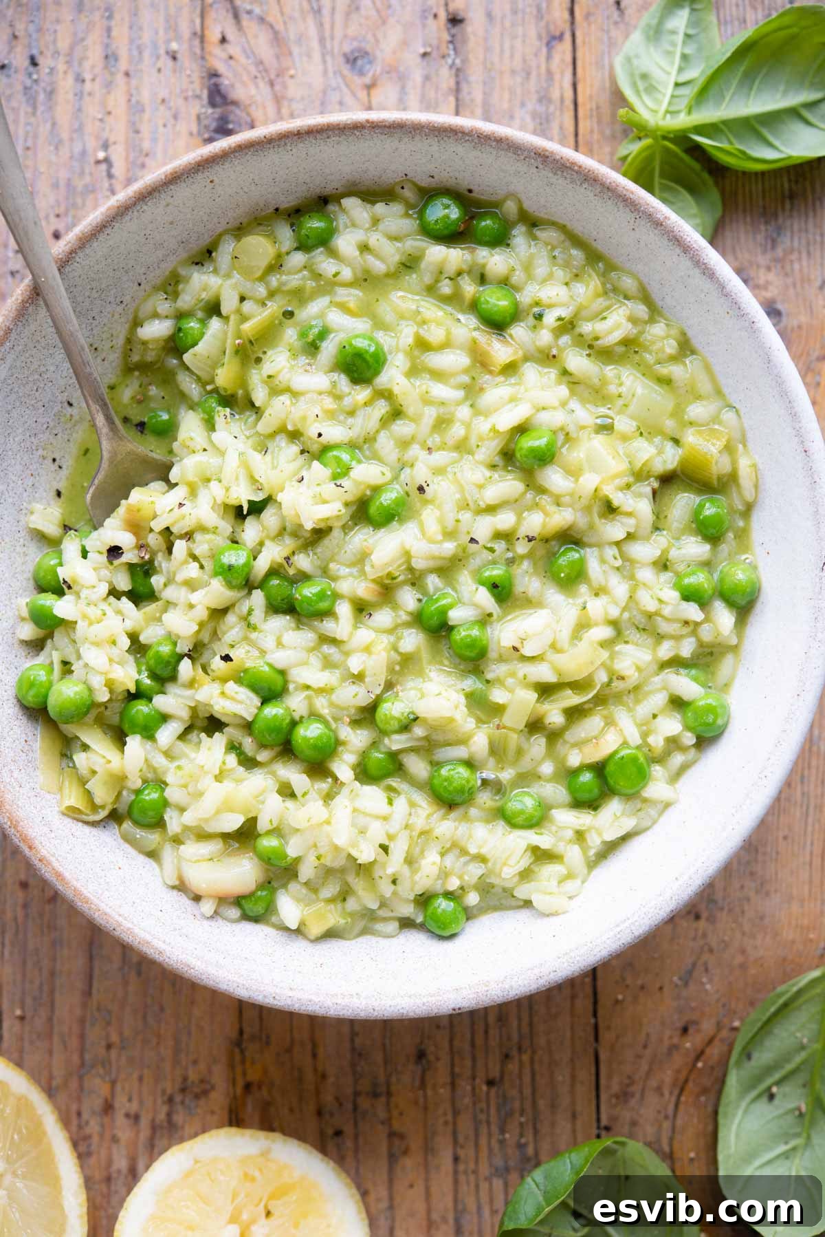 An overhead shot of a bowl of creamy pea and pesto risotto garnished with fresh basil leaves and lemon slices on a rustic wooden background.