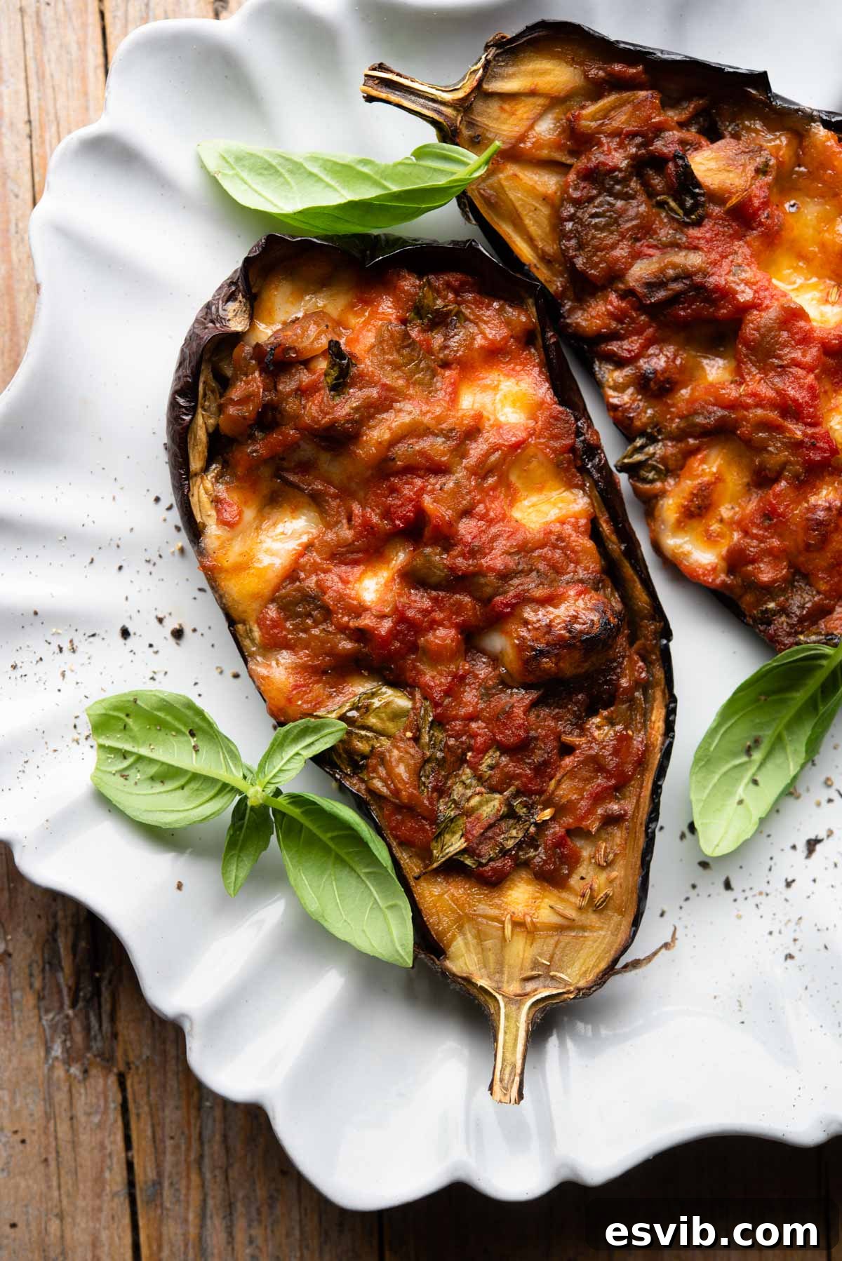 Two perfectly baked Italian stuffed eggplants, topped with melted mozzarella and vibrant fresh basil, artfully arranged on a rustic scalloped-edged plate.