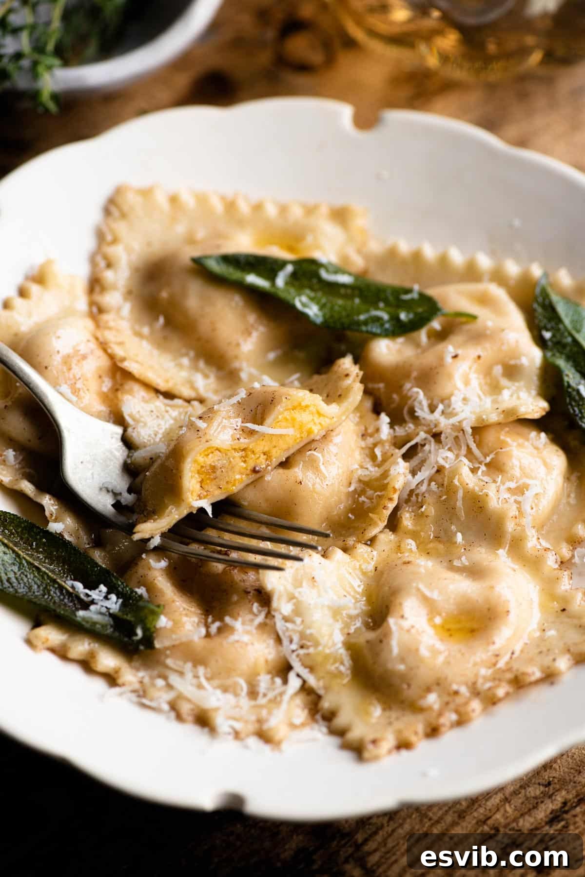 Golden Butternut Ravioli with Aromatic Sage Brown Butter 11 A close-up, appetizing shot of golden butternut squash ravioli nestled in a bowl with glistening sage brown butter sauce. One ravioli is cut in half, revealing the vibrant orange, creamy filling within, garnished with fresh sage.