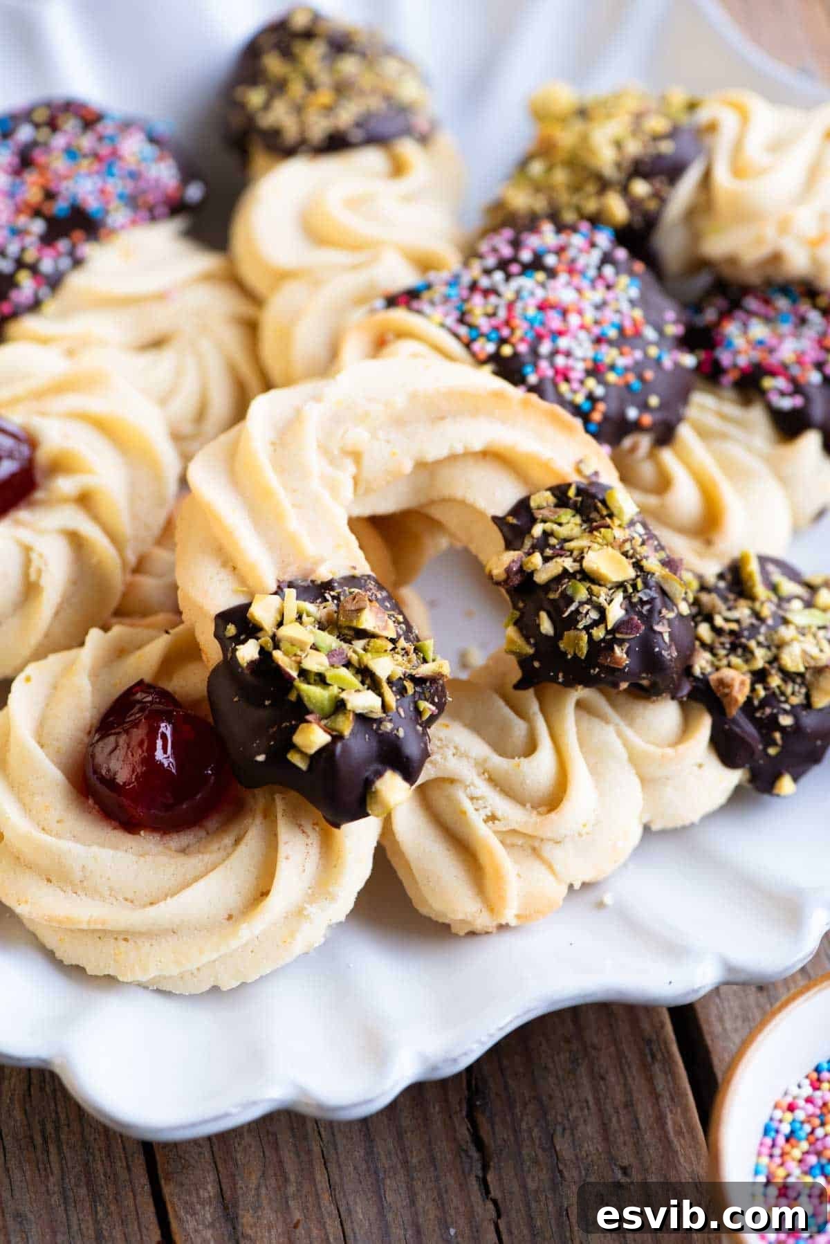 Italian Whipped Butter Cookies 7 A captivating close-up of elegantly presented Italian butter cookies on a serving plate. Some are partially dipped in dark chocolate and generously sprinkled with finely chopped pistachios, highlighting their artisanal charm.