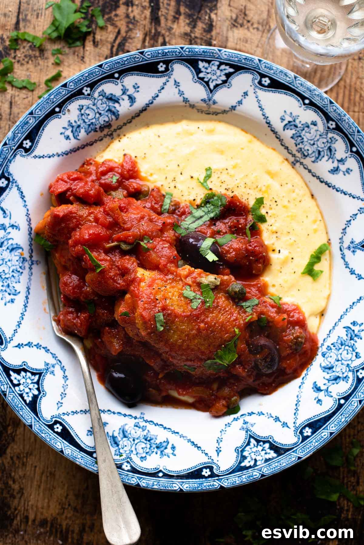 Chicken in Puttanesca sauce in a bowl with creamy polenta sitting on a rustic wooden surface, garnished with fresh parsley.