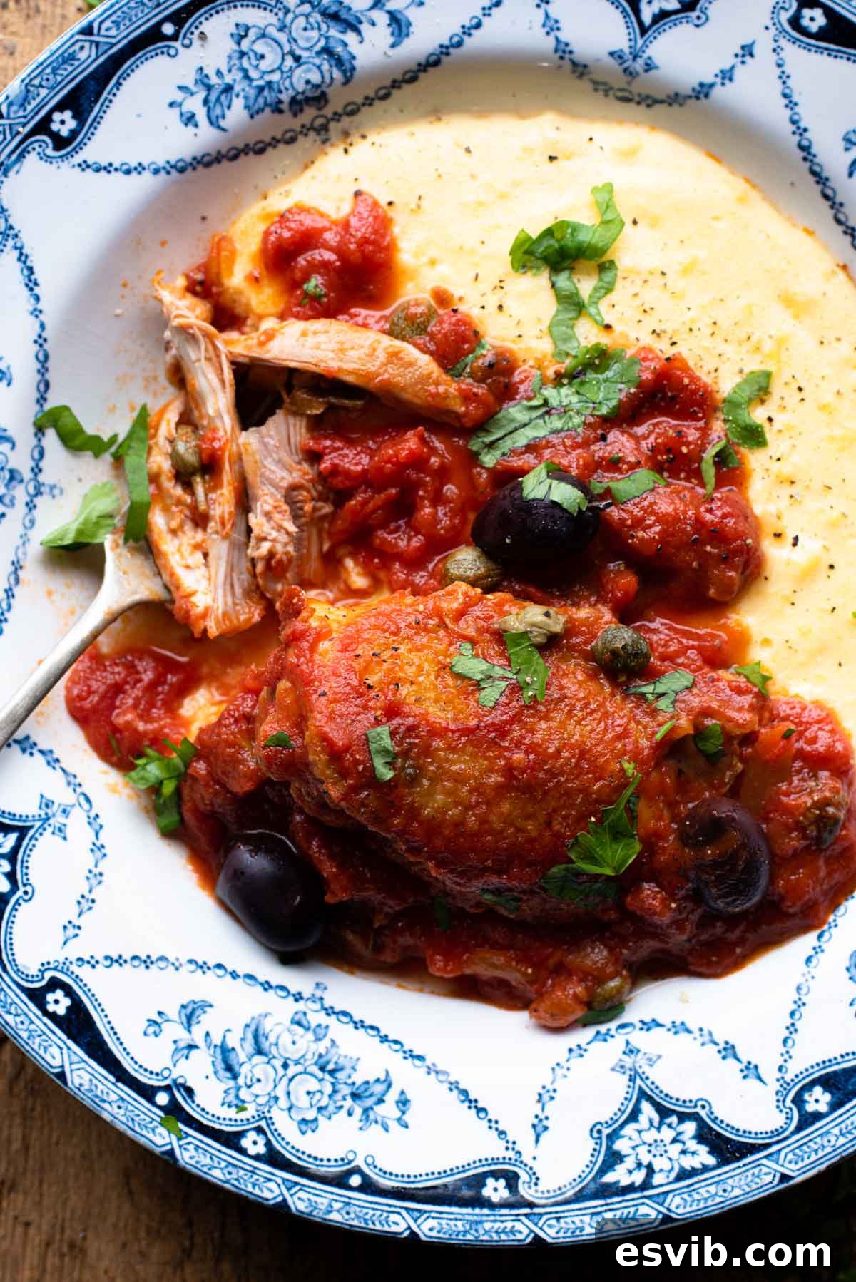 A close-up shot of Chicken Puttanesca served with creamy polenta in a bowl, placed on a wooden table, emphasizing the rich texture of the sauce and the tender chicken.