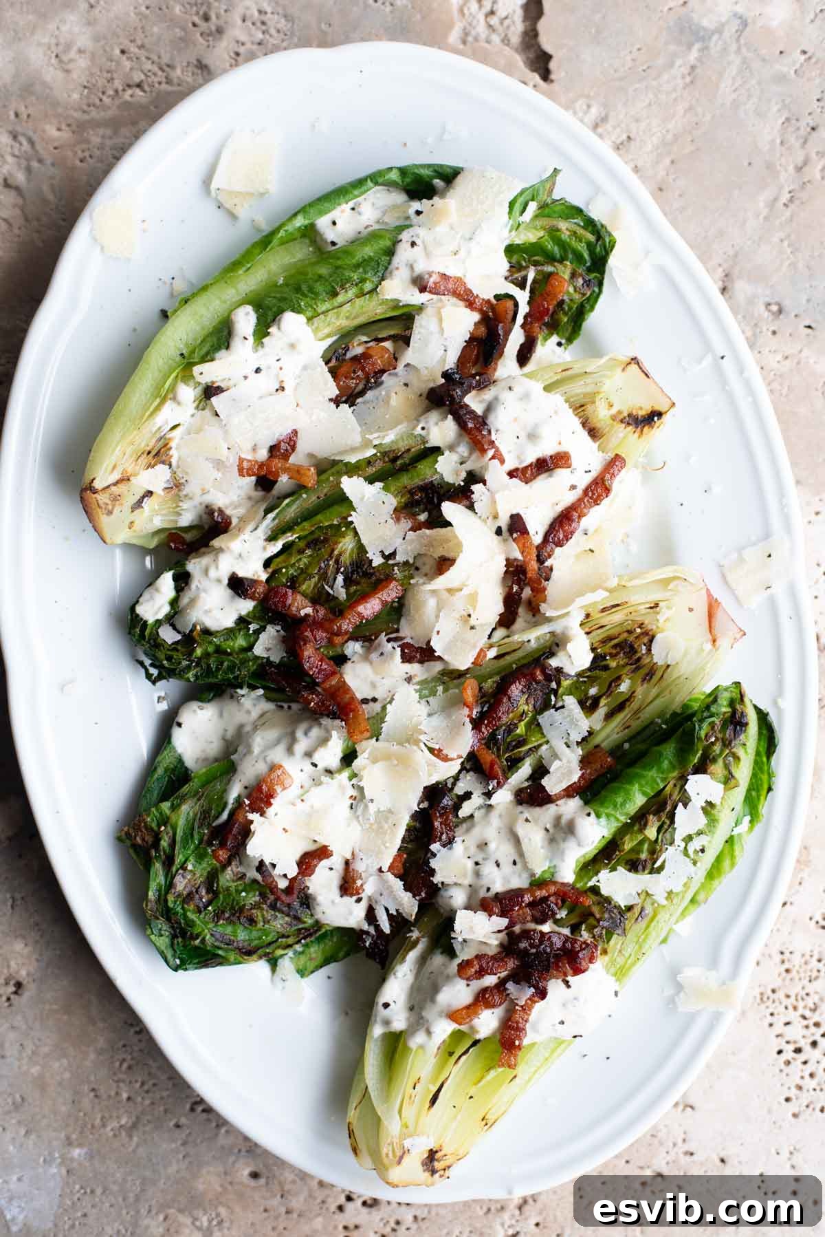 An overhead shot of a white serving plate with grilled romaine lettuce topped with a creamy sauce, pancetta and Parmigiano Reggiano shavings.