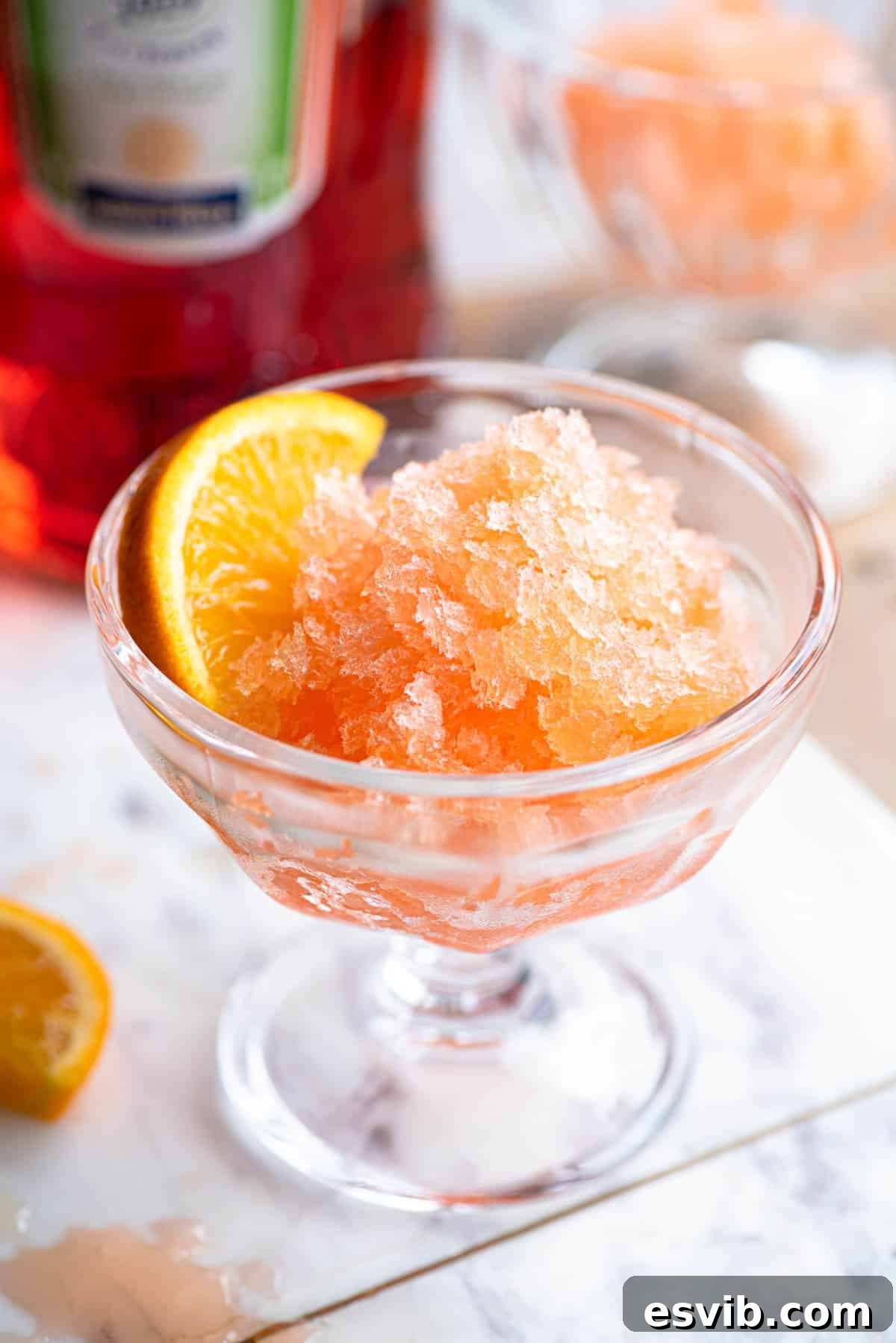 Aperol granita in a small glass bowl with a slice of orange at the side sitting on a marble board.