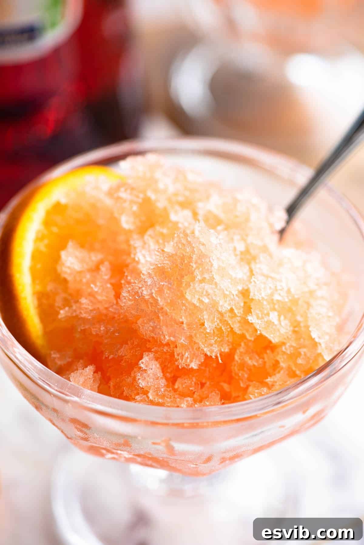 A close up of Aperol granita in a small glass bowl with a spoon and slice of orange at the side.