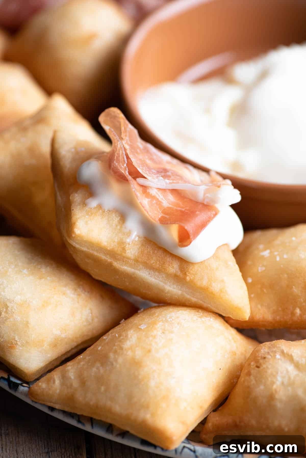 A vibrant close-up shot of crispy Gnocco Fritto, perfectly puffed and golden, artfully arranged on a plate with fresh slices of cheese and prosciutto atop, inviting indulgence.