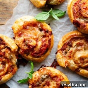 A close up overhead shot of golden-brown pizza pinwheels arranged on a sheet of baking parchment, garnished with fresh basil leaves, highlighting their flaky texture.
