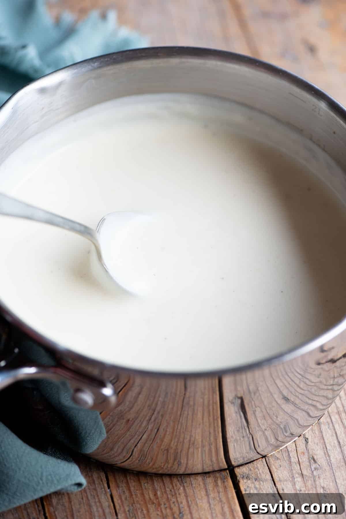 Classic Italian Béchamel Sauce 2 Silky smooth Italian Besciamella sauce in a saucepan, with a spoon dipping into it, on a rustic wooden background.