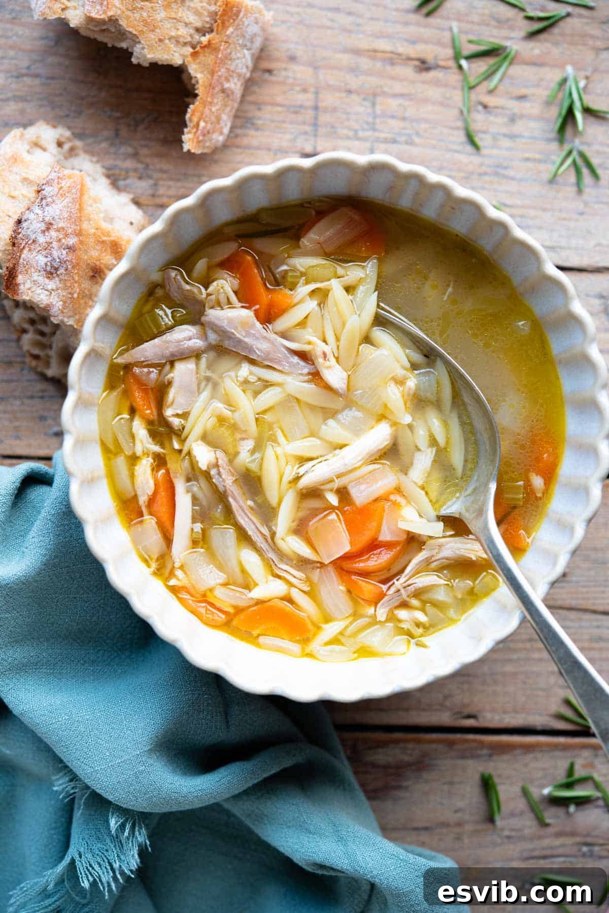 An inviting overhead shot of a bowl of vibrant Lemon Chicken Orzo Soup, garnished with fresh herbs, resting on a rustic wooden surface. A slice of crusty bread sits enticingly beside it.