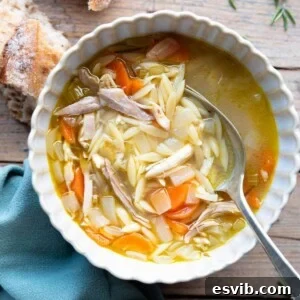 An inviting overhead shot of lemon chicken orzo soup in a bowl. The background features a wooden surface with crusty bread at the side, evoking a cozy meal.