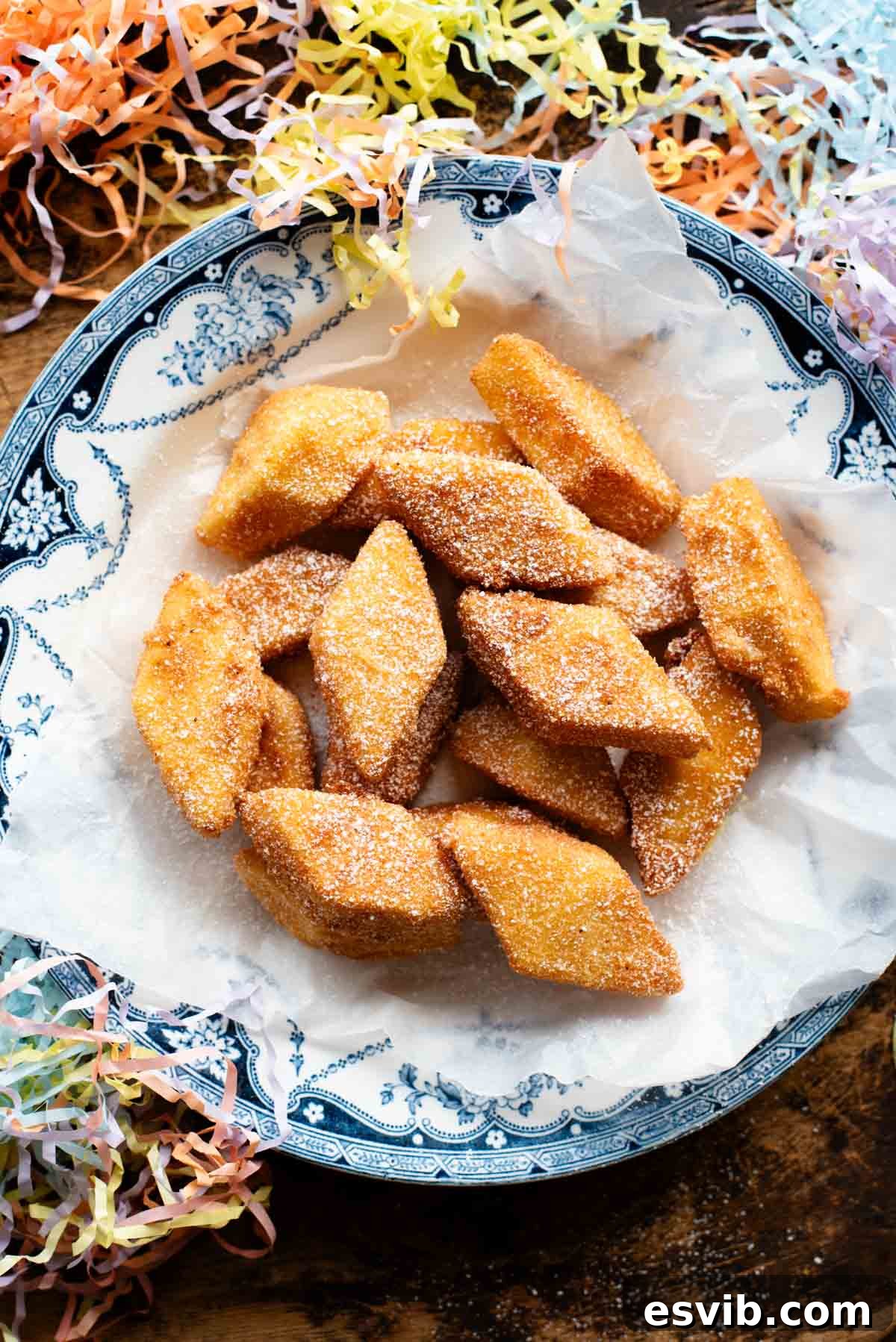 An overhead shot of golden-brown Crema Fritta (fried custard bites) arranged in a decorative bowl, heavily topped with powdered sugar. Festive confetti is playfully scattered around the bowl, suggesting a celebration.