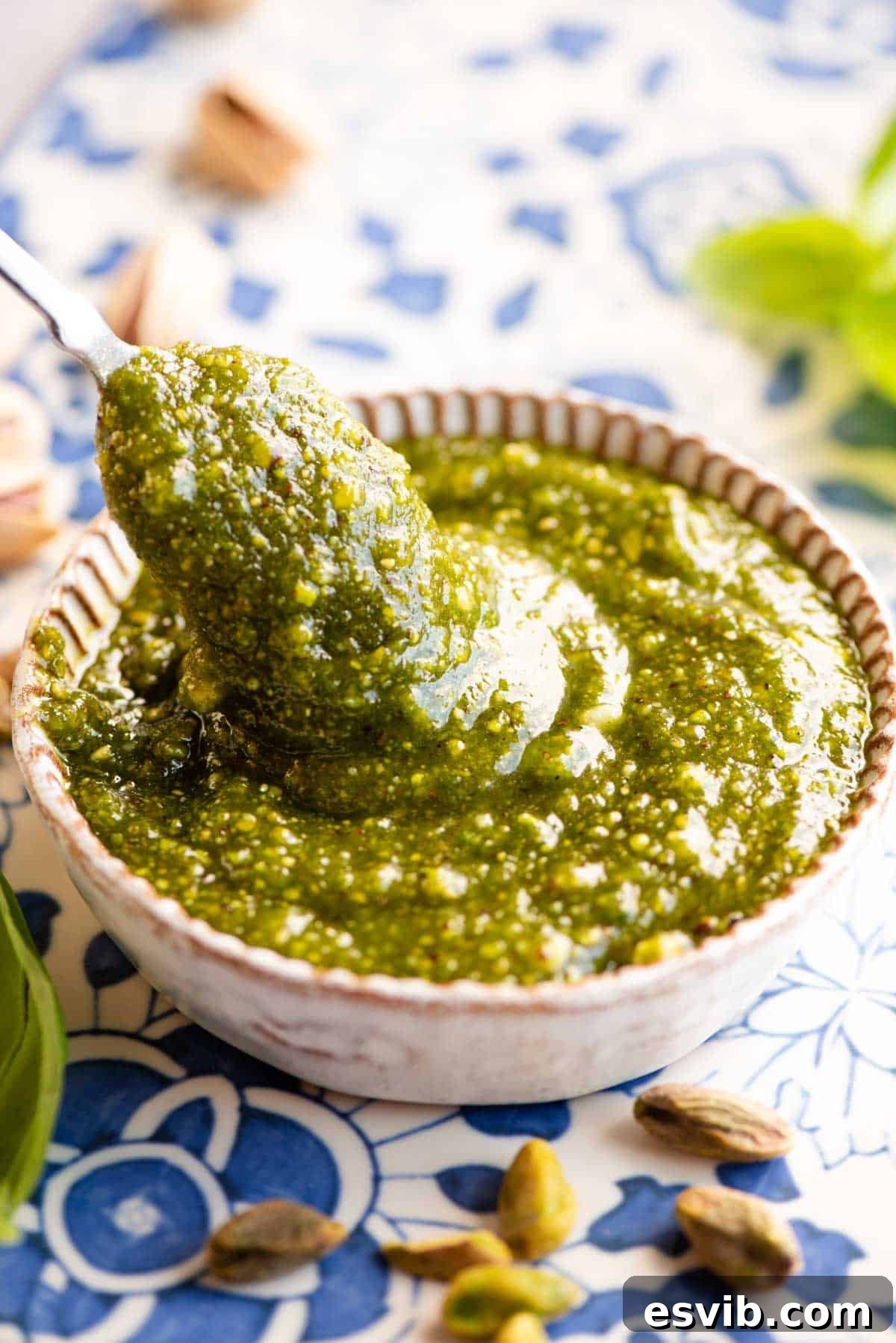 A close up of a spoon in a dish of pistachio pesto sitting on a blue patterned background with pistachios around. The texture of the pesto is visible.