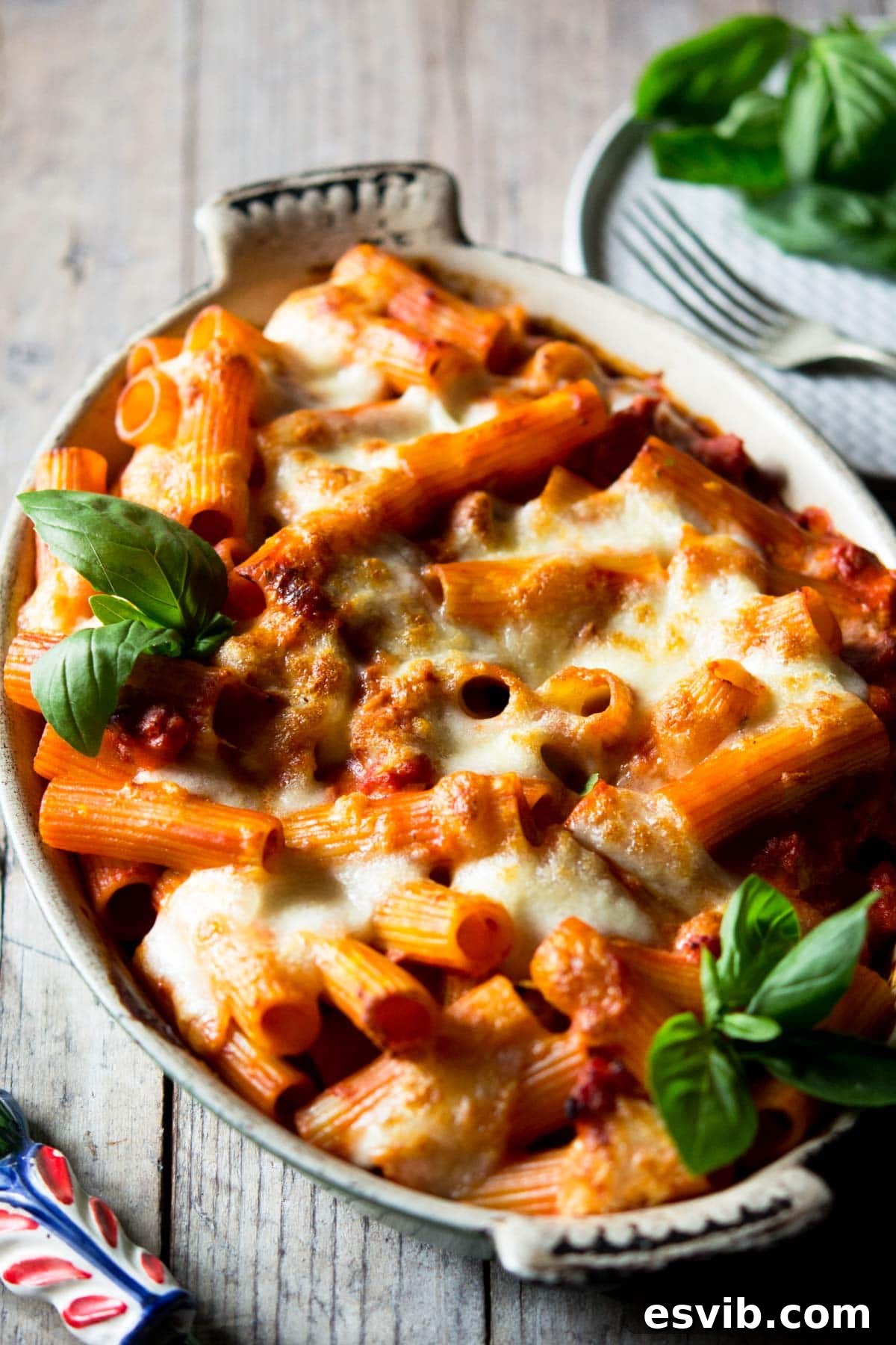 Sausage pasta bake made with rigatoni pasta in an oval baking dish with melted mozzarella A close-up of a bubbling Sausage Pasta Bake in a casserole dish, garnished with fresh basil.