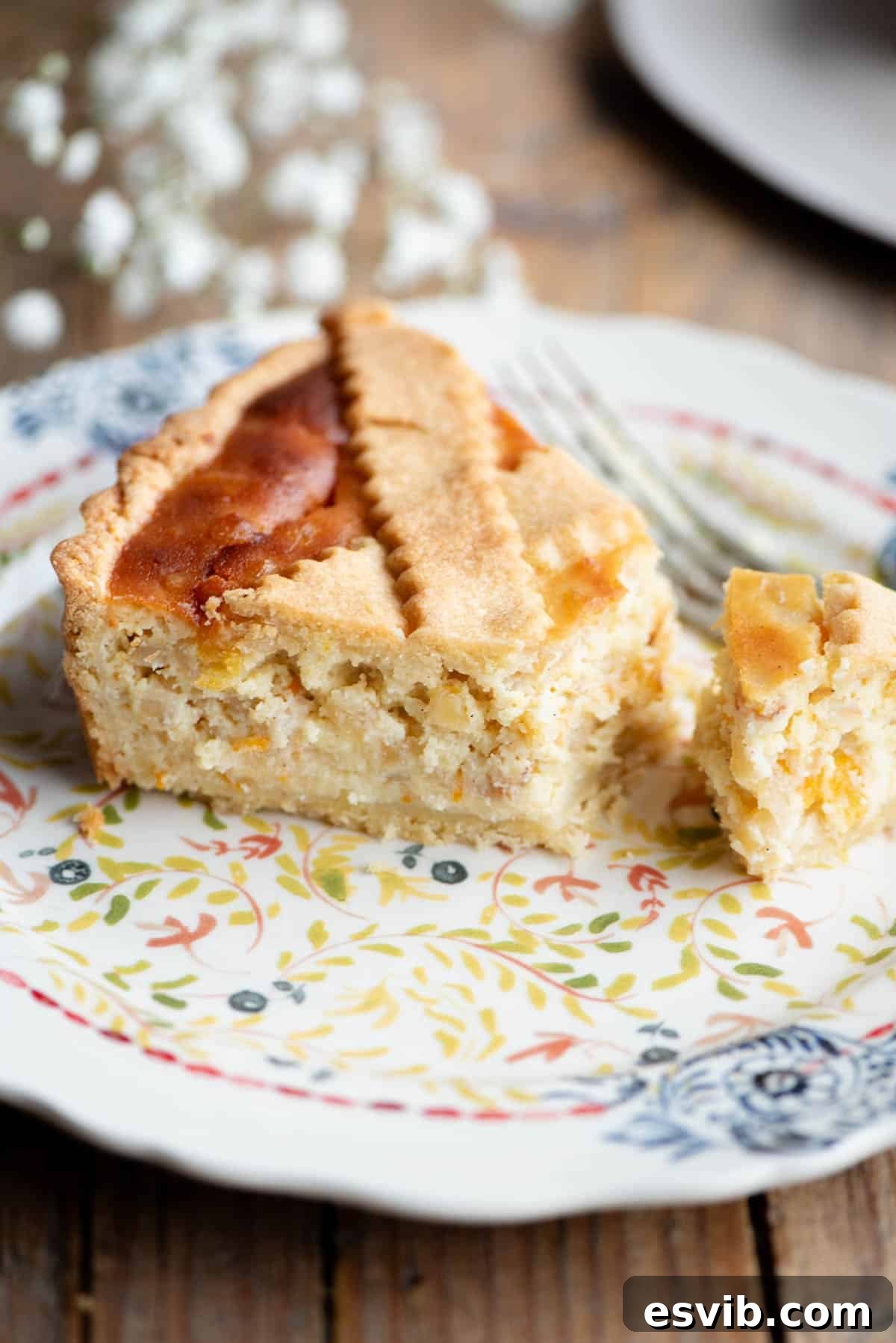 A perfect slice of Pastiera Napoletana, revealing its rich, creamy filling and delicate crust, served on a patterned plate with soft white flowers artfully blurred in the background.
