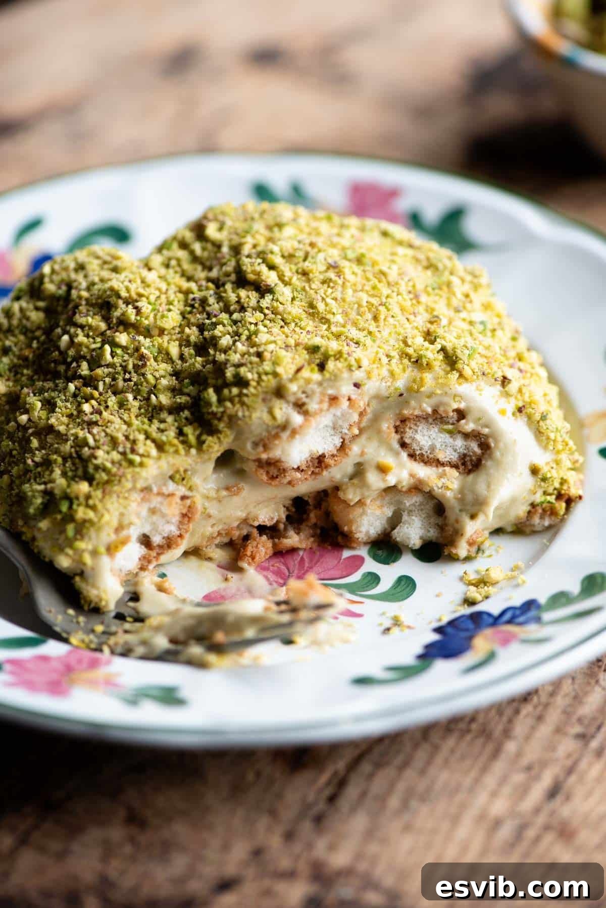 A close up of a piece of pistachio tiramisu on a plate sitting on a wooden table, showcasing its creamy layers and pistachio crumb topping.