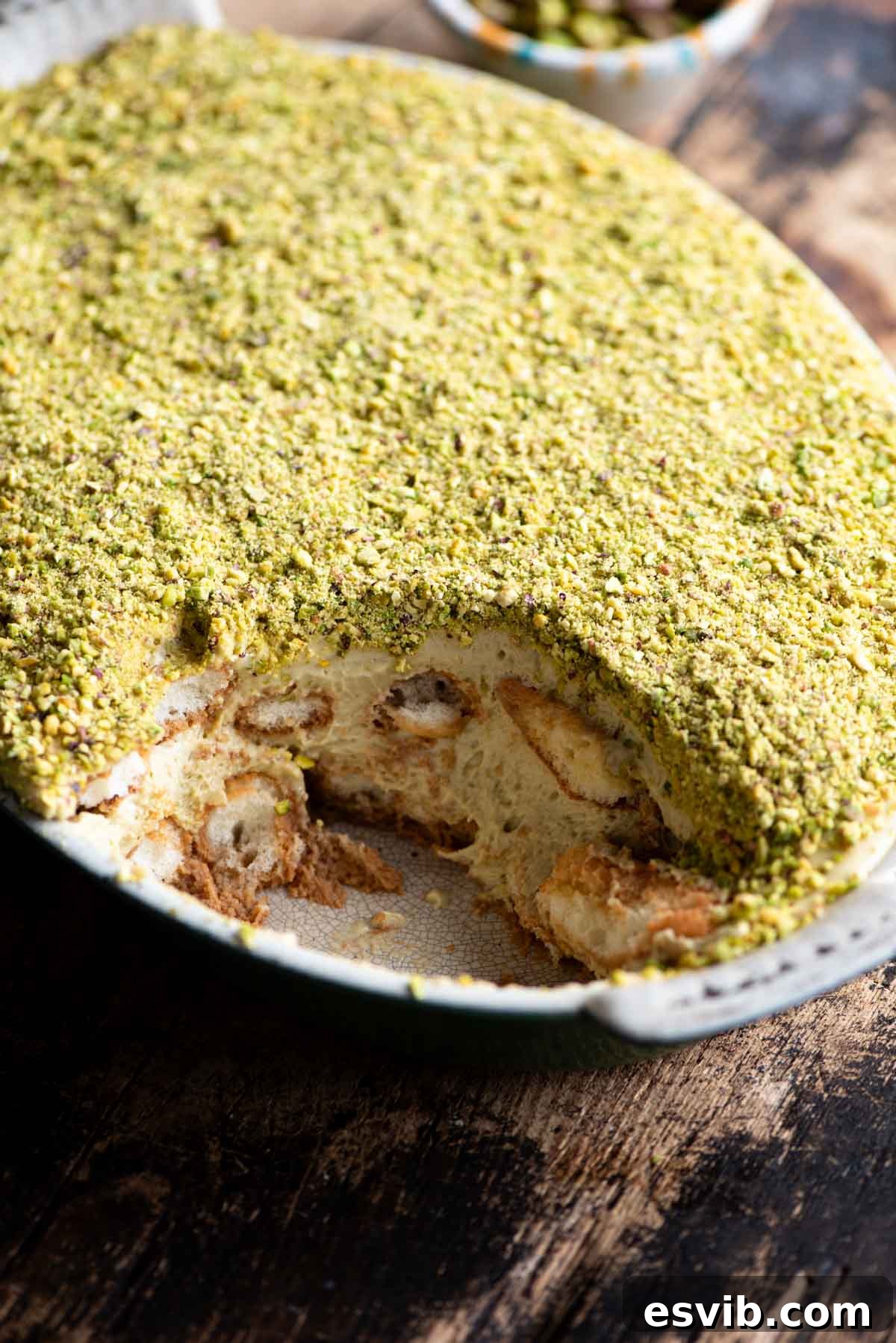 A side shot of a pistachio tiramisu in an oval dish with a slice removed, revealing the delicious layers. The background is wooden and rustic.