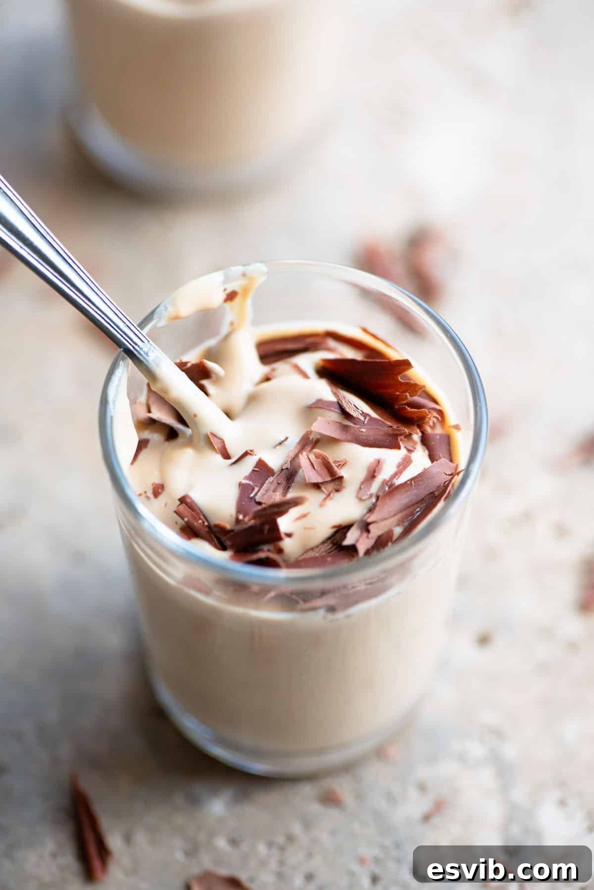 A close-up view of Ricotta al Caffè served in a small clear glass, with a spoon resting inside. The texture is creamy and smooth, and subtle specks of dark chocolate are visible.
