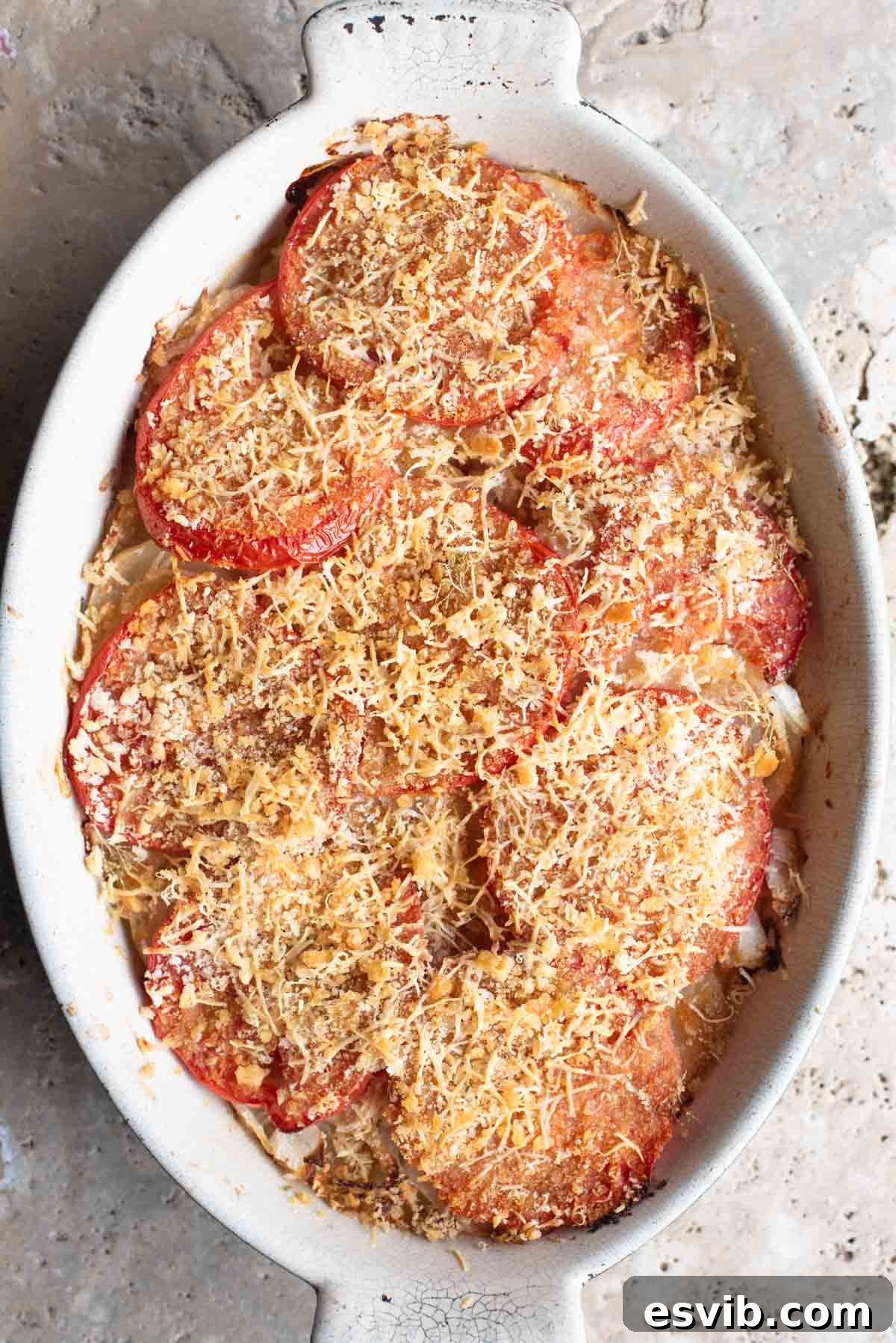 An overhead shot of an Italian potato and tomato gratin in an oval dish sitting on a Travertine background. The potatoes and tomatoes are perfectly baked with a crispy breadcrumb topping.