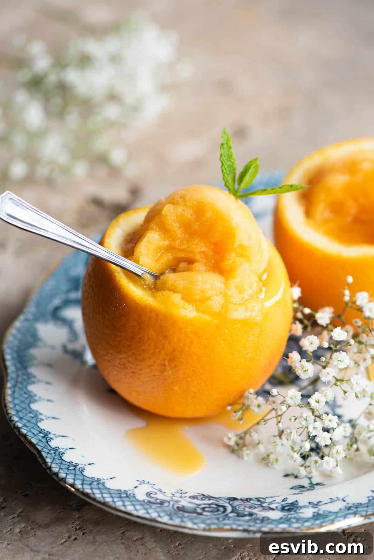 A close-up shot of refreshing orange sorbet served beautifully inside a hollowed-out orange peel, resting on a blue plate alongside delicate flowers.