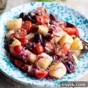 A close-up cropped image of a Sicilian potato salad in a blue bowl with olives, onions and tomatoes.