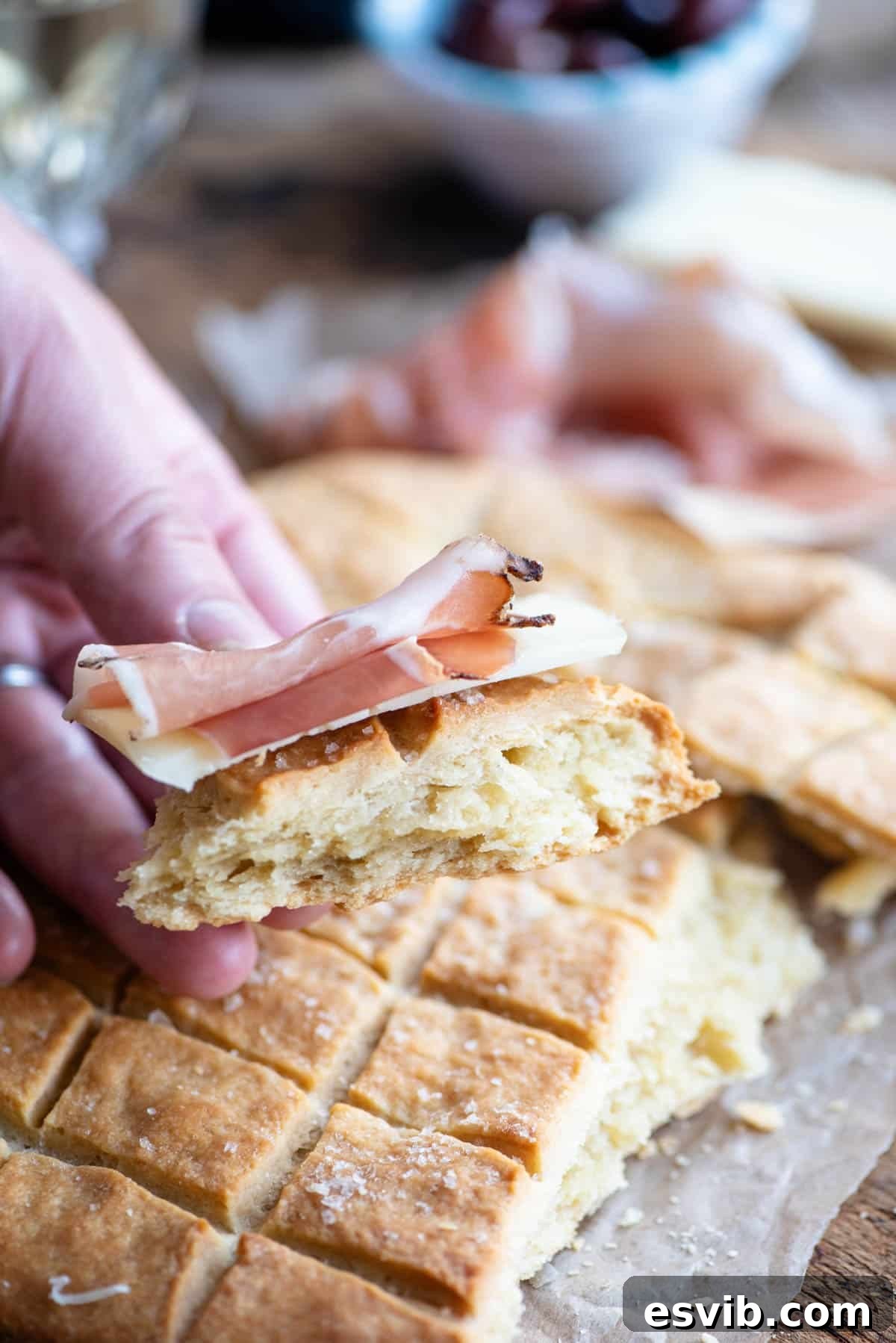A close-up shot of a hand gracefully holding a piece of golden-brown Pizza Scima, with a sliver of cured meat delicately draped over it, highlighting its perfect crispness.