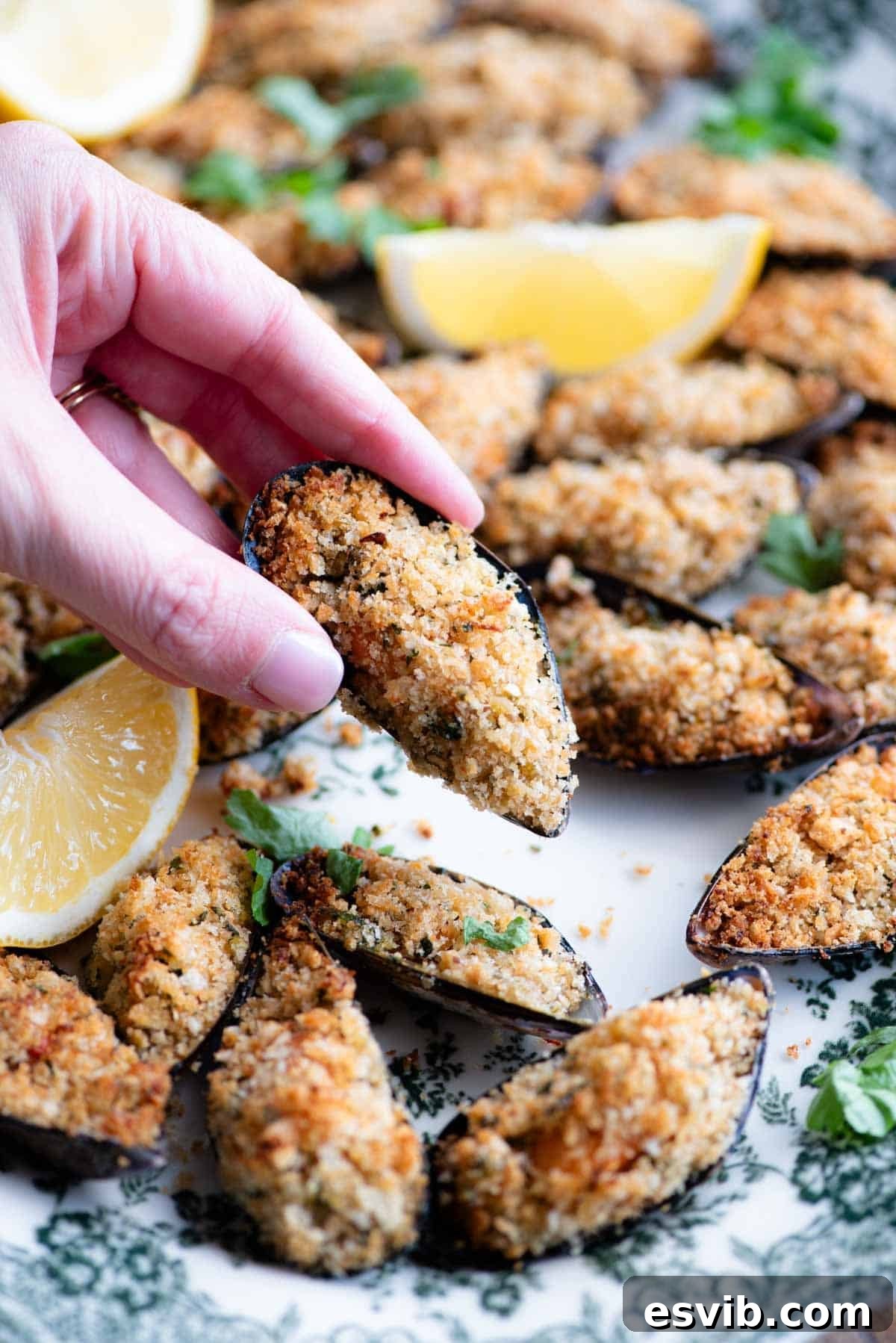 A close-up shot of a hand gently holding a perfectly baked Italian stuffed mussel, poised over a decorative serving plate, highlighting its golden crust.