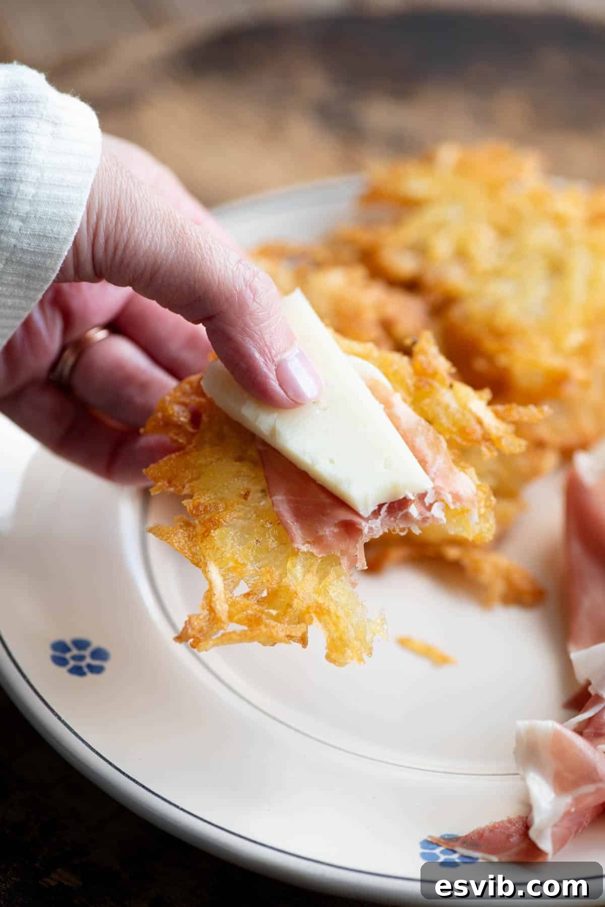 A close-up shot of a hand gracefully holding a single, perfectly crispy Tortei di Patate, adorned with delicate slices of cured meat and a piece of creamy cheese, highlighting its deliciousness as a savory bite.