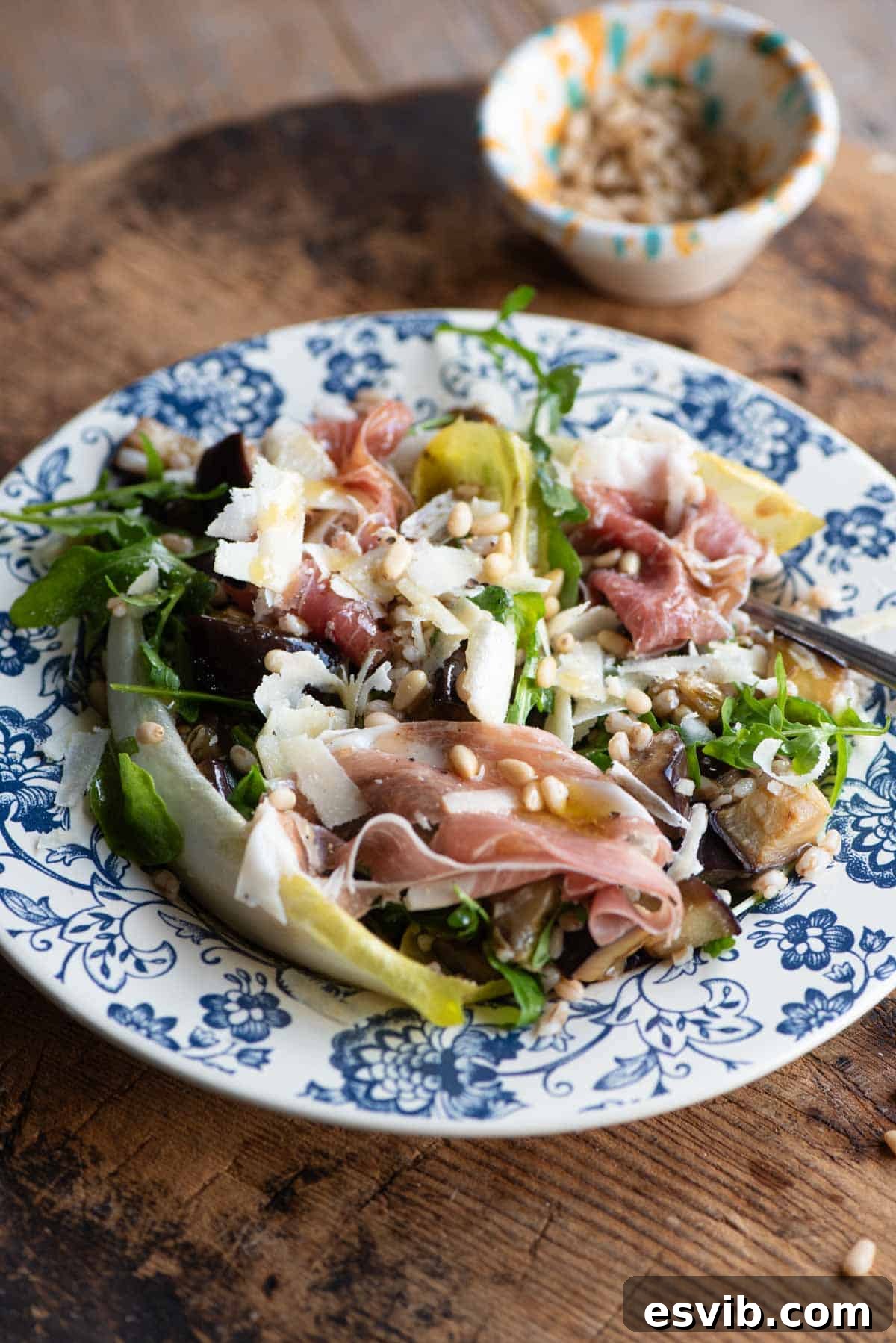 Italian Harvest Farro with Prosciutto 2 A close up of an eggplant and farro salad on a blue patterned plate on a wooden table, garnished with prosciutto and pine nuts.