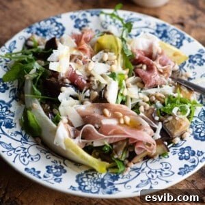 A close up cropped image of an eggplant and farro salad on a blue plate with prosciutto and pine nuts, ready to be enjoyed.
