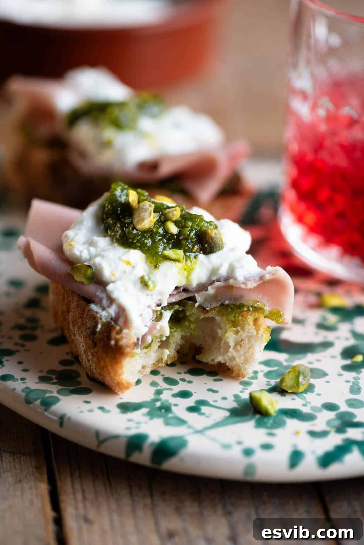 Pistachio-Crusted Mortadella and Burrata Crostini 4 A close up of a single mortadella, burrata and pistachio crostini with a bite taken out, showing the creamy interior and crispy bread.