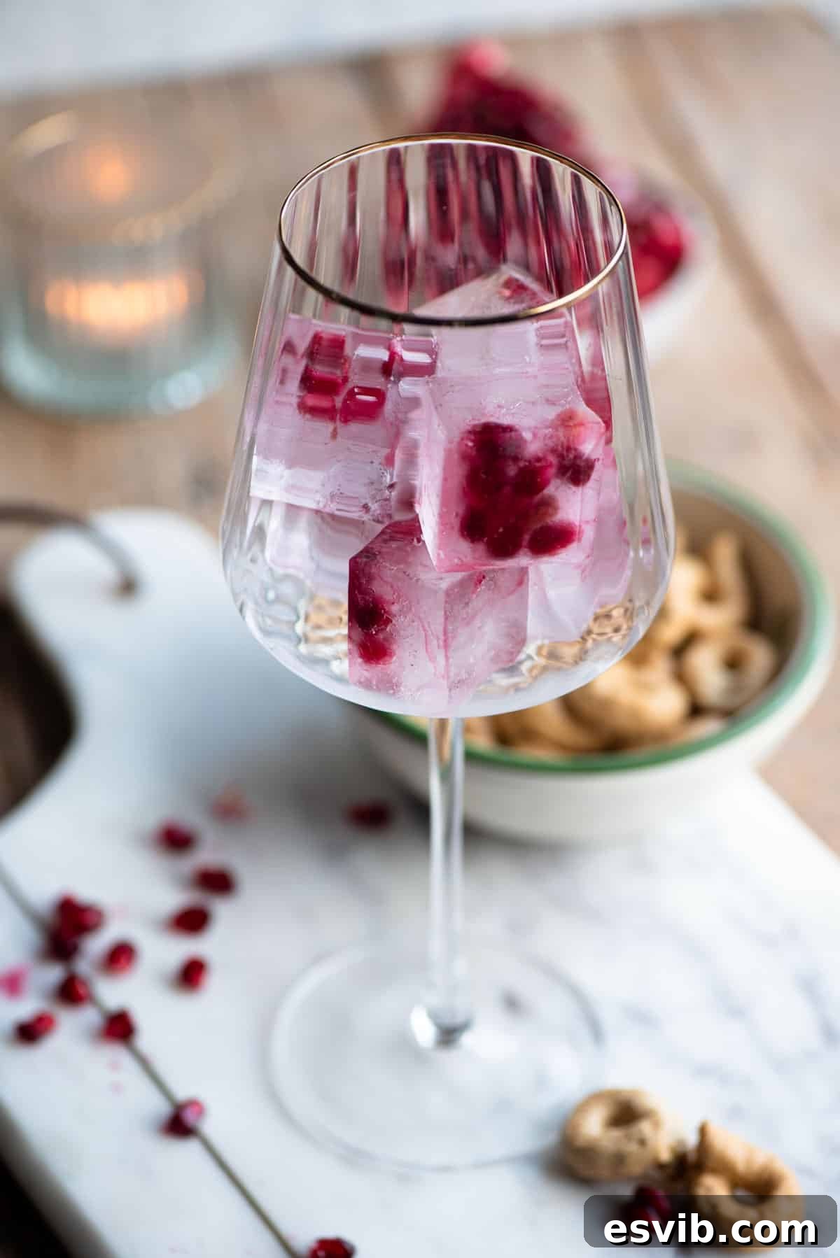 Crimson Sparkler 3 A close-up shot of a sophisticated wine glass with a delicate gold rim, filled with custom-made pomegranate ice cubes. The vibrant red arils suspended within the ice create a visually stunning and festive appearance, ready to be infused into a cocktail.
