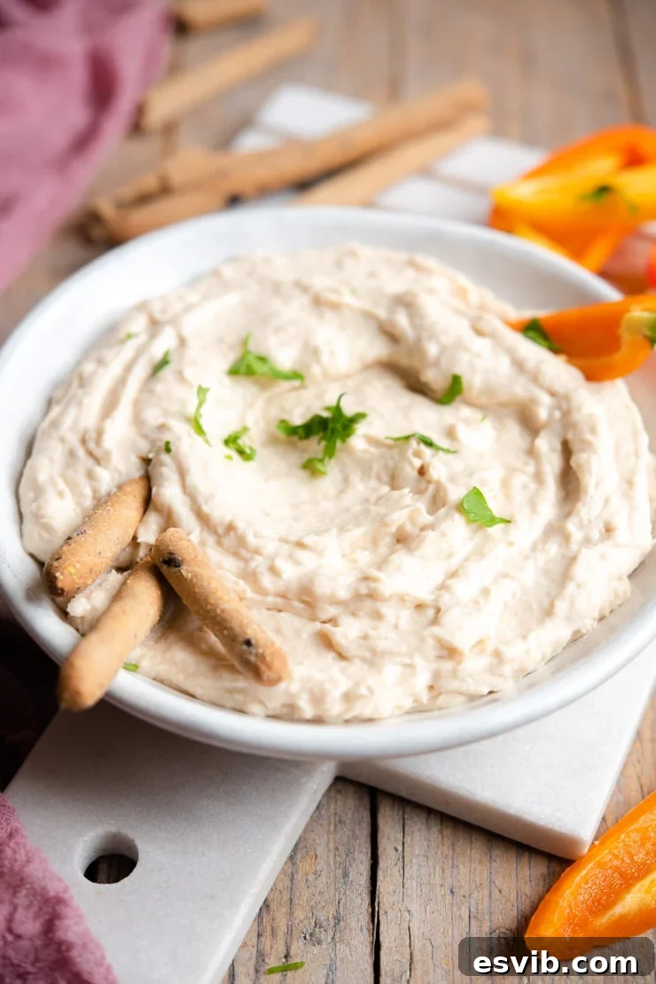A side shot of a creamy white bean dip in a ceramic bowl, artfully arranged with golden breadsticks and vibrant red bell pepper slices for dipping.