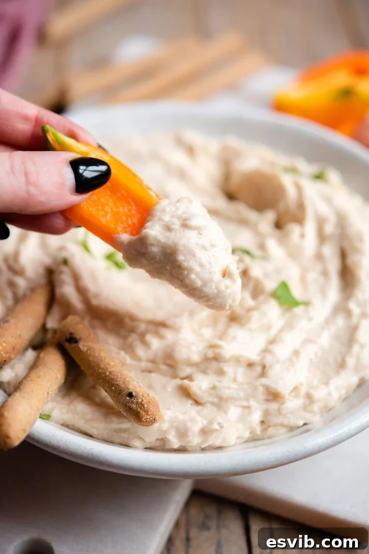A close-up shot of a hand dipping a vibrant red bell pepper slice into a bowl of creamy white bean dip, highlighting its smooth texture.