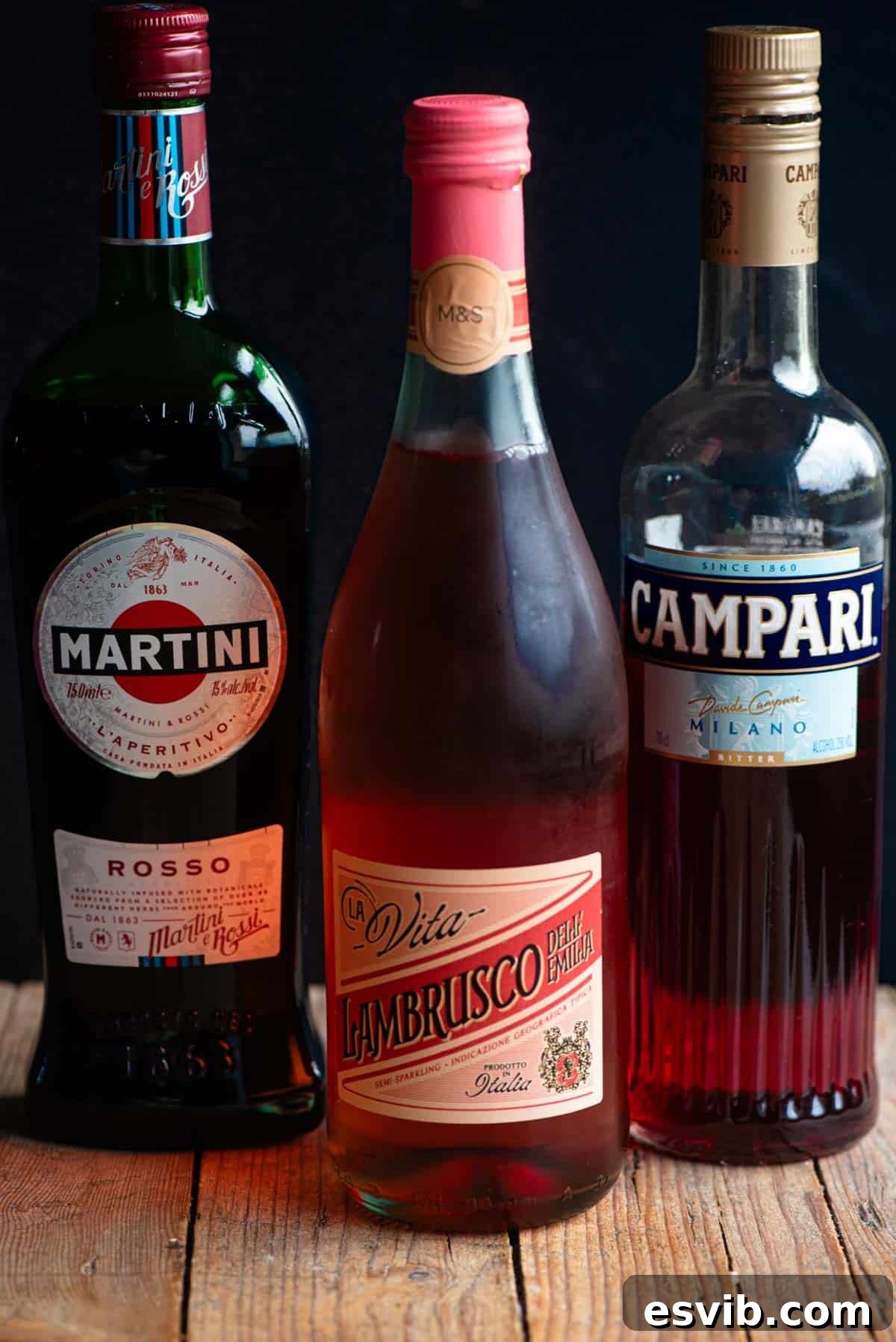 Scarlet Serendipity 3 A rustic wooden table displaying a bottle of Martini Rosso, a bottle of sparkling Lambrusco, and a bottle of Campari, all essential ingredients for the Sbagliato Rosso, set against a dark, moody background.
