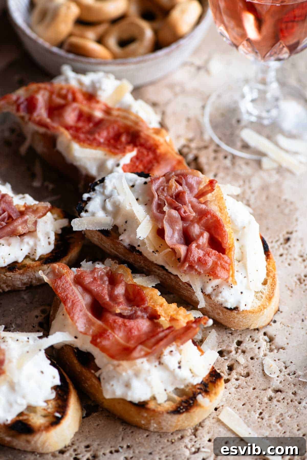 A close up of a perfectly assembled slice of crostini topped generously with creamy burrata, grated Parmigiano Reggiano, and a piece of crispy prosciutto.