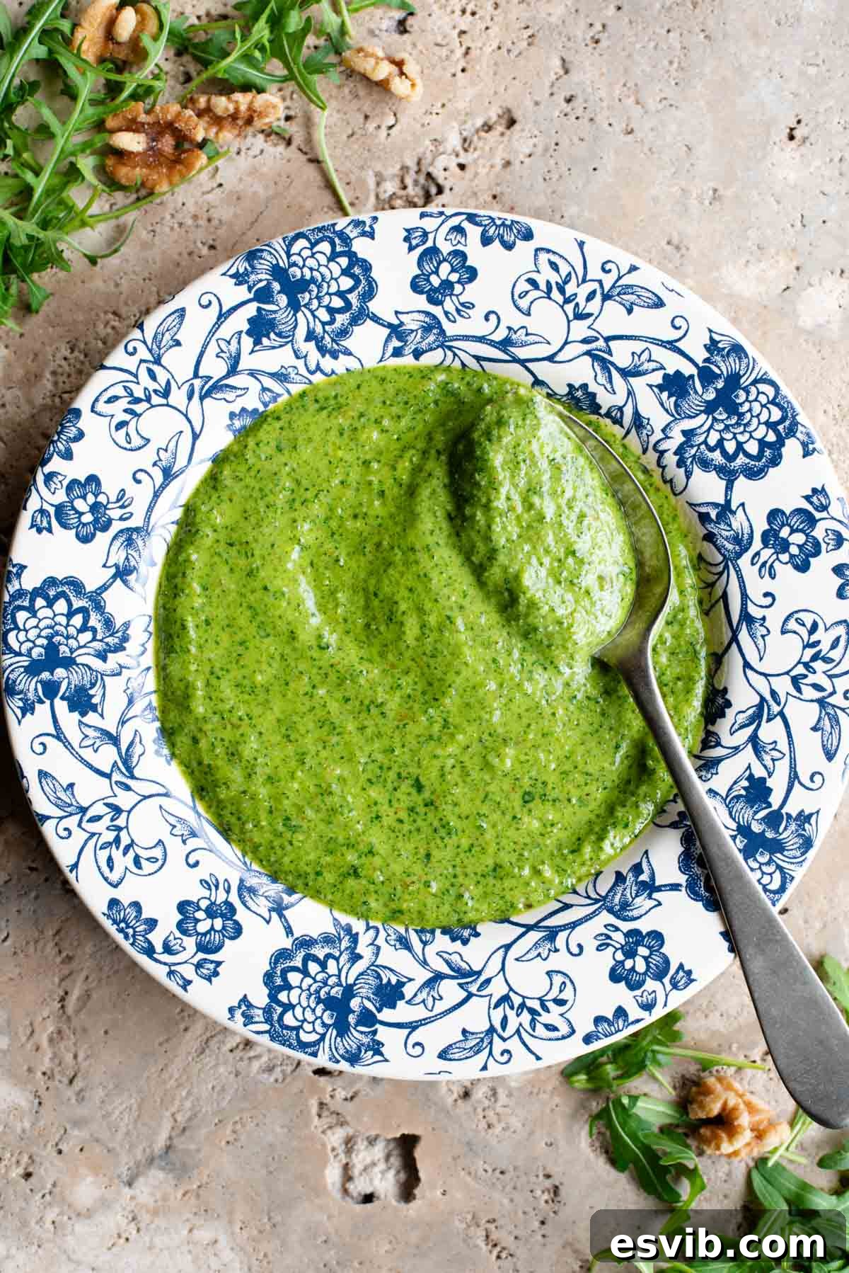 An overhead shot of vibrant green arugula pesto in a beautiful blue and white patterned bowl with a silver spoon resting beside it, ready to be enjoyed.
