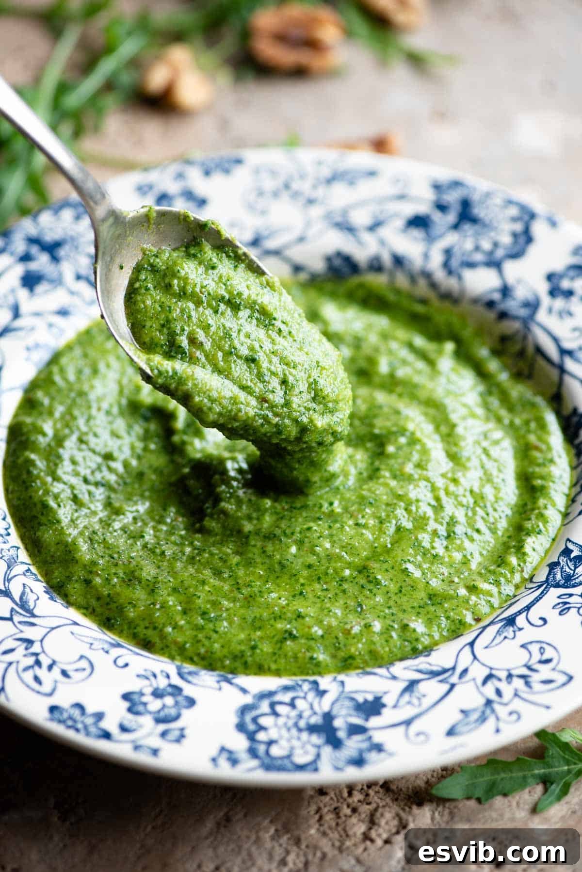 A close up shot of a spoon gently scooping up some freshly made arugula pesto from a vibrant blue and white patterned bowl, showcasing its rich texture.