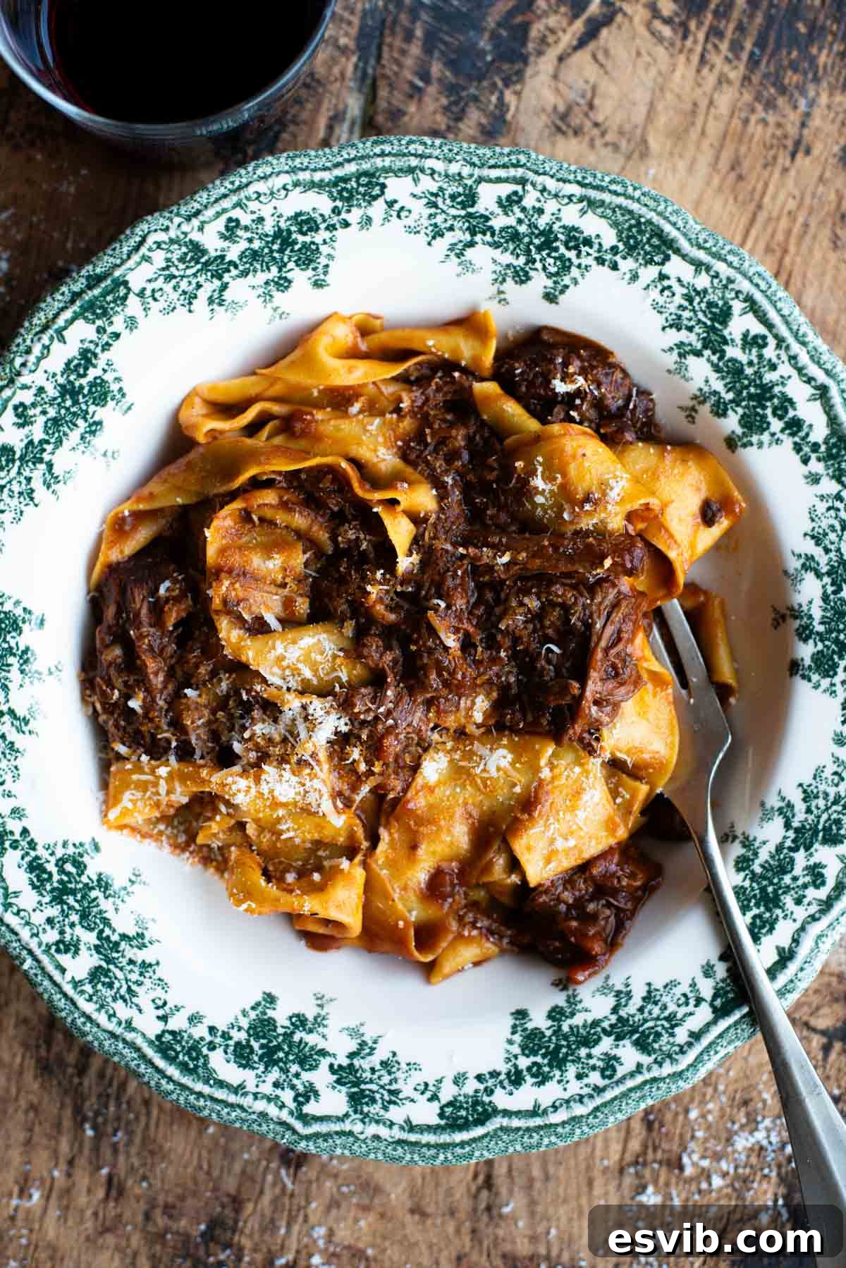 An overhead shot of a beef short rib ragu, glistening with rich sauce, served in a vibrant green and white bowl with wide pappardelle pasta strands, ready to be enjoyed.