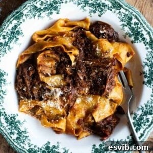 A close up of beef rib ragu with pappardelle pasta in a green and white bowl, garnished with fresh Parmigiano Reggiano.