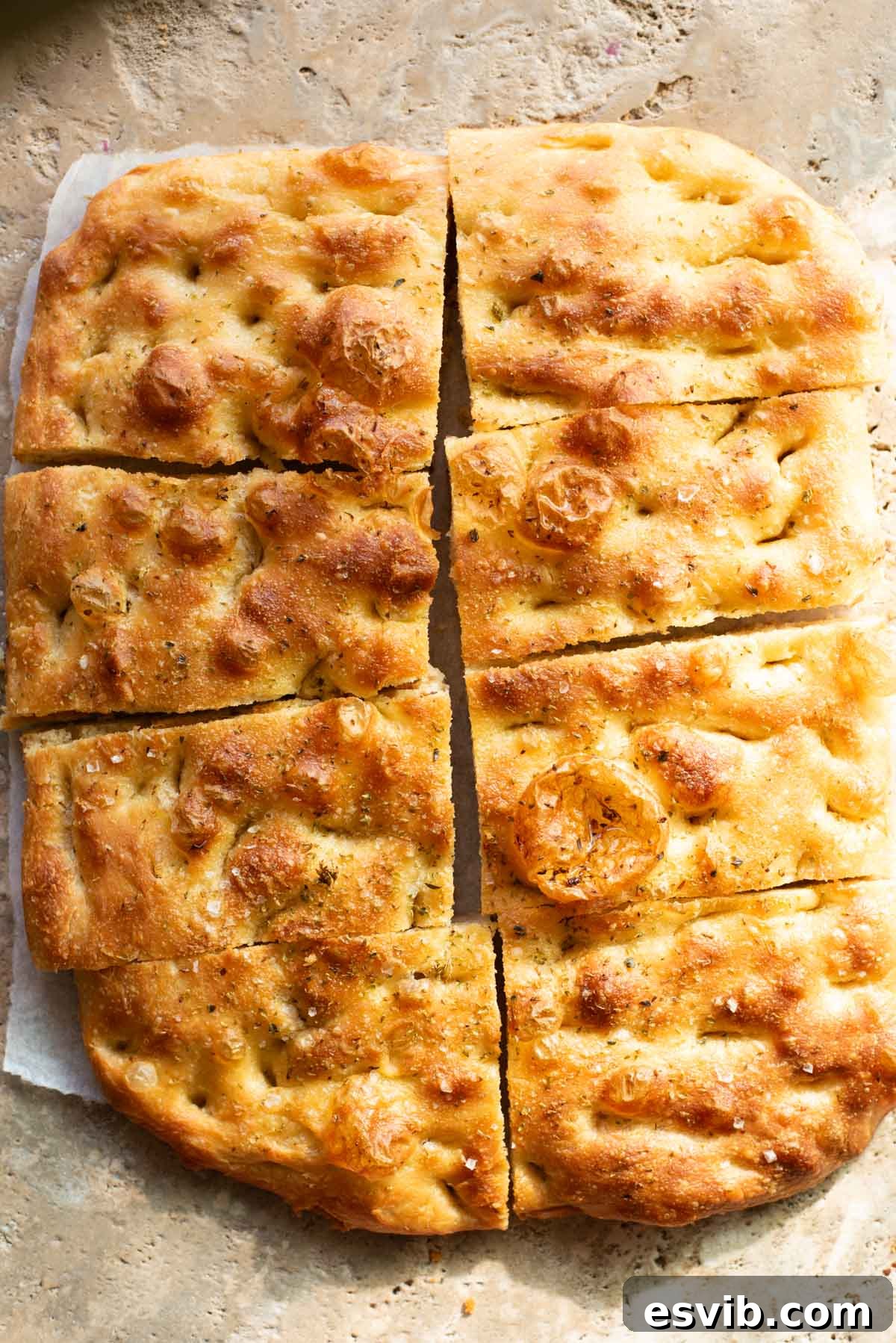 Golden, rustic focaccia bread sliced into rectangular pieces, with a crisp, dimpled crust, a sprinkle of sea salt, and a light seasoning of herbs, resting on a stone surface.