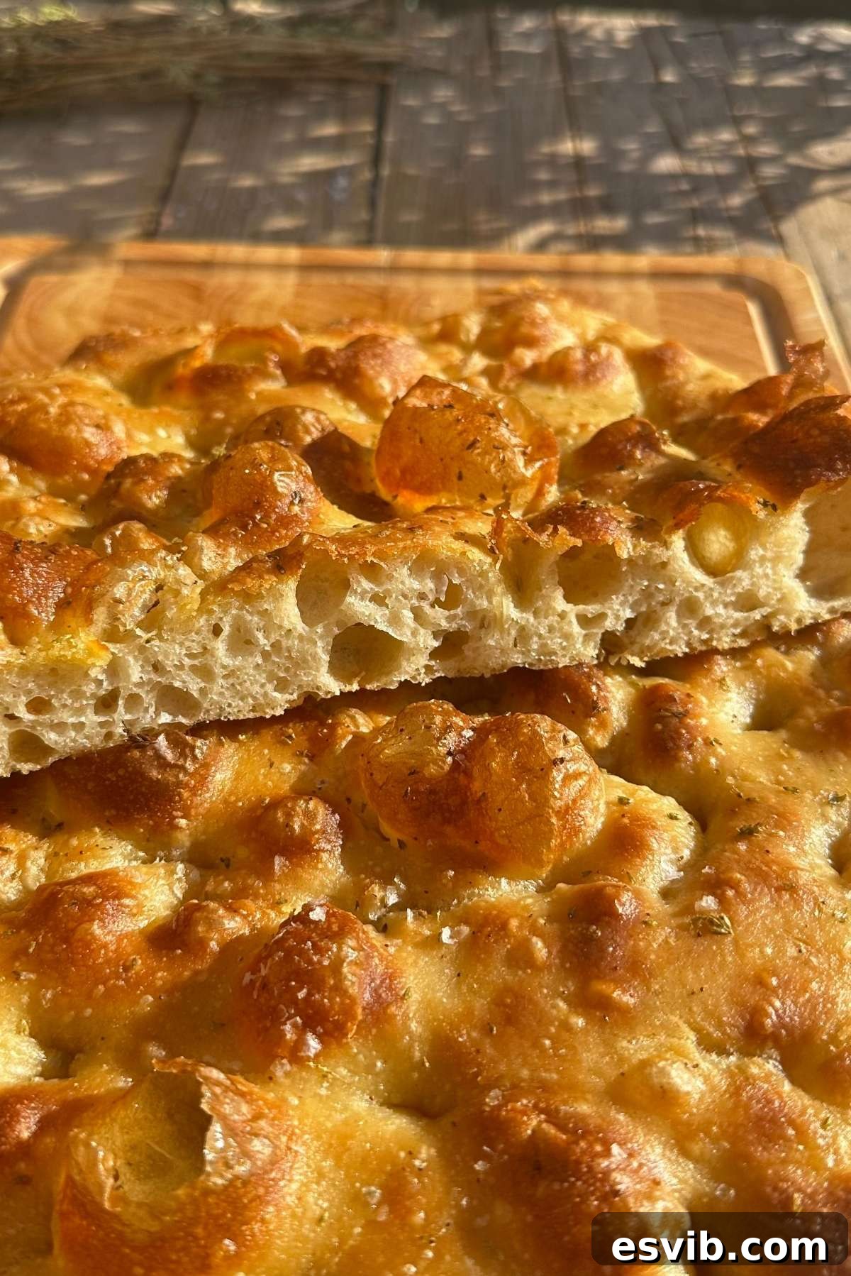 A golden and airy focaccia cut in half to show it's bubbly texture sitting on a wooden chopping board.