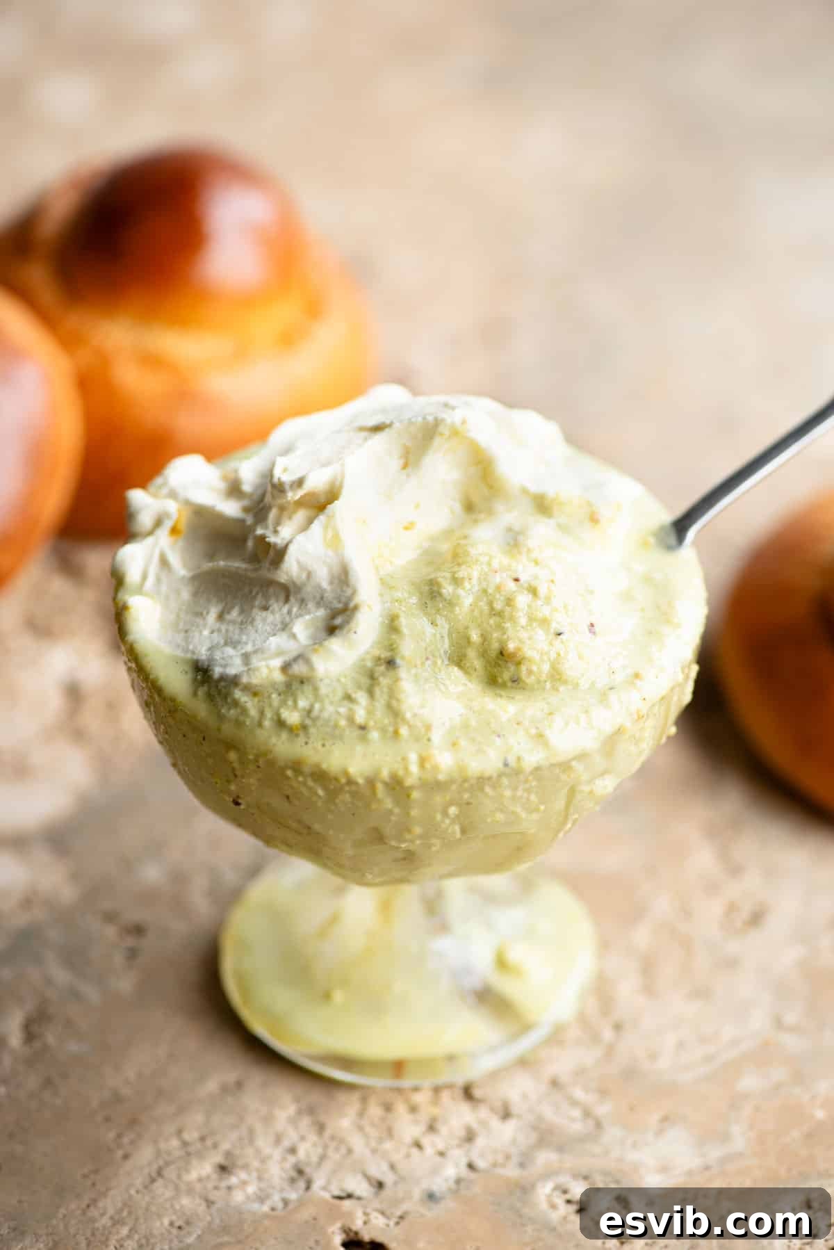 A close up of pistachio granita in a glass serving bowl topped with whipped cream and a brioche bun behind it.