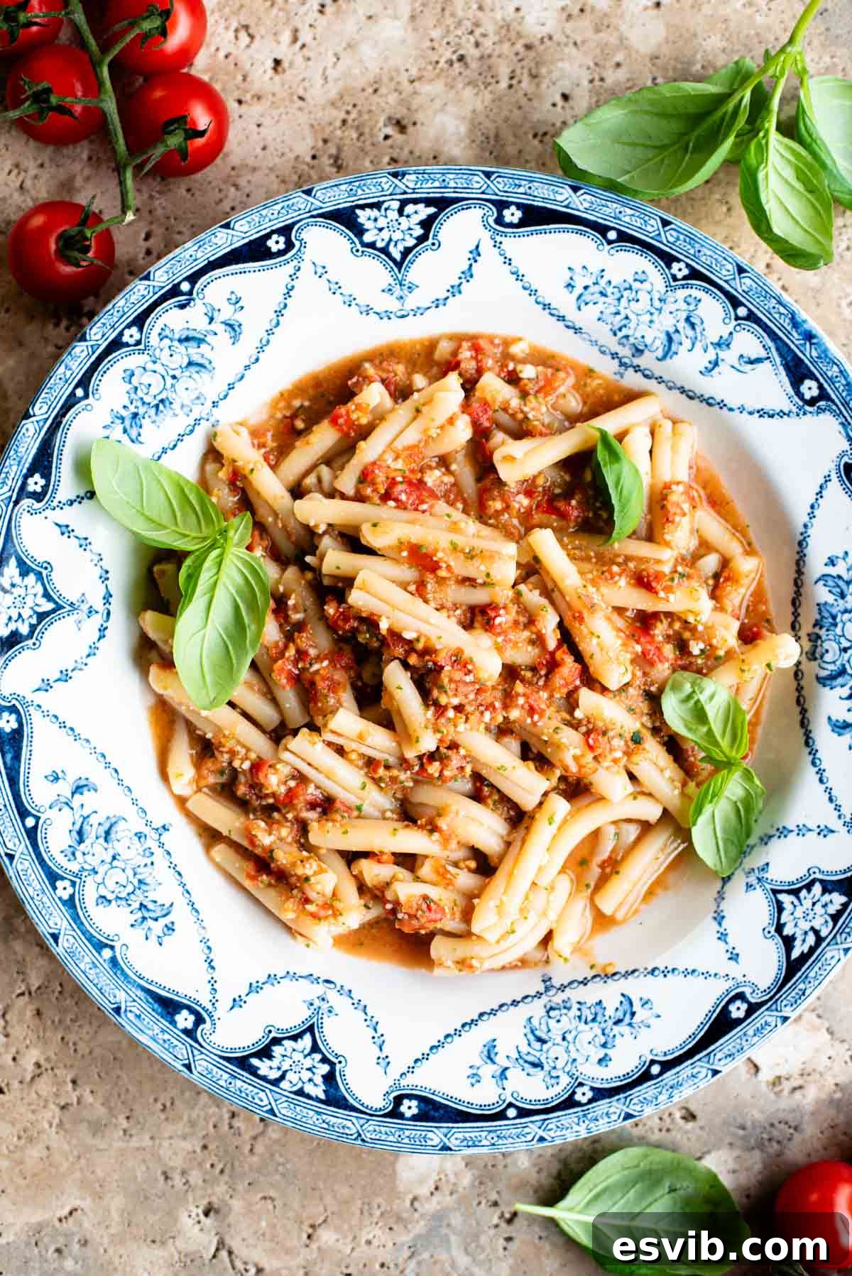 A blue and white vintage bowl with pasta in a fresh tomato sauce topped with basil. The bowl is on a travertine surface with cherry tomatoes and basil around.
