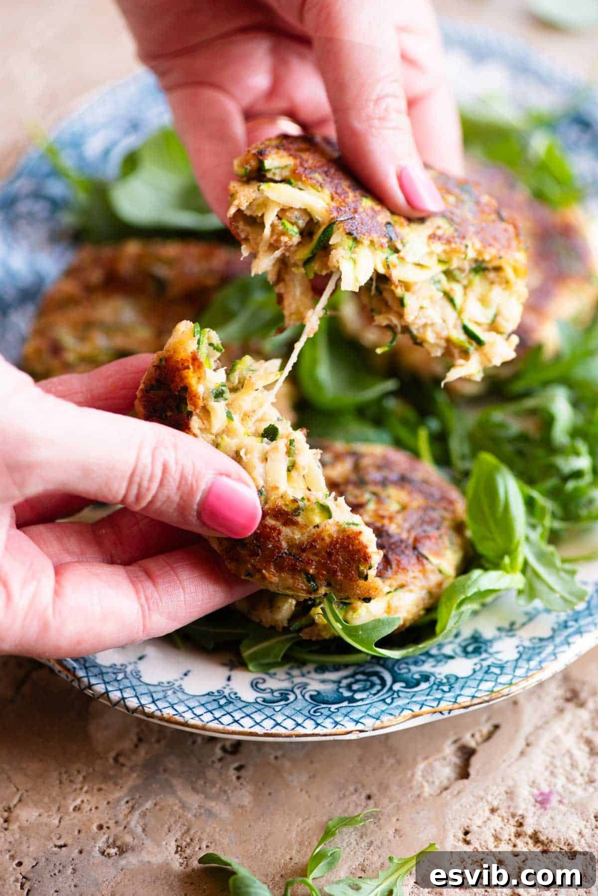 Two hands gently pulling apart a cooked zucchini patty, showcasing the melted, stretchy cheese creating a tempting cheese pull in the middle, highlighting its delicious texture.