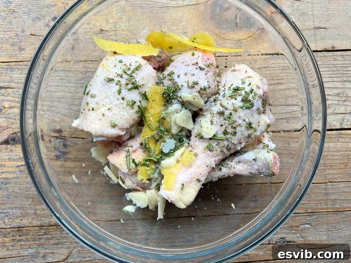 Chicken thighs and drumsticks marinating in a glass bowl with lemon zest, fresh herbs, and garlic, positioned on a rustic wooden surface.