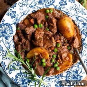 A close-up of a bubbling beef stew with tender potatoes and vibrant green peas, garnished with fresh rosemary, served in a charming blue and white bowl.