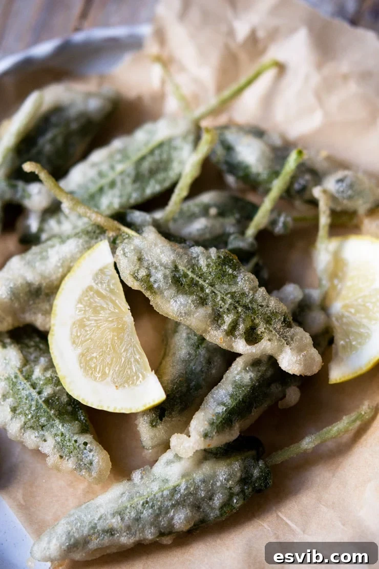 A close-up view of golden-brown fried sage leaves with anchovies, neatly piled on a white plate, garnished with fresh lemon wedges, highlighting their irresistible crisp texture.