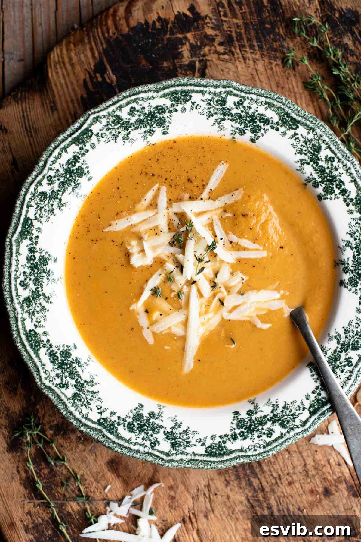 An overhead shot of a bowl of roasted vegetable soup in a green and white vintage bowl on a wooden board.