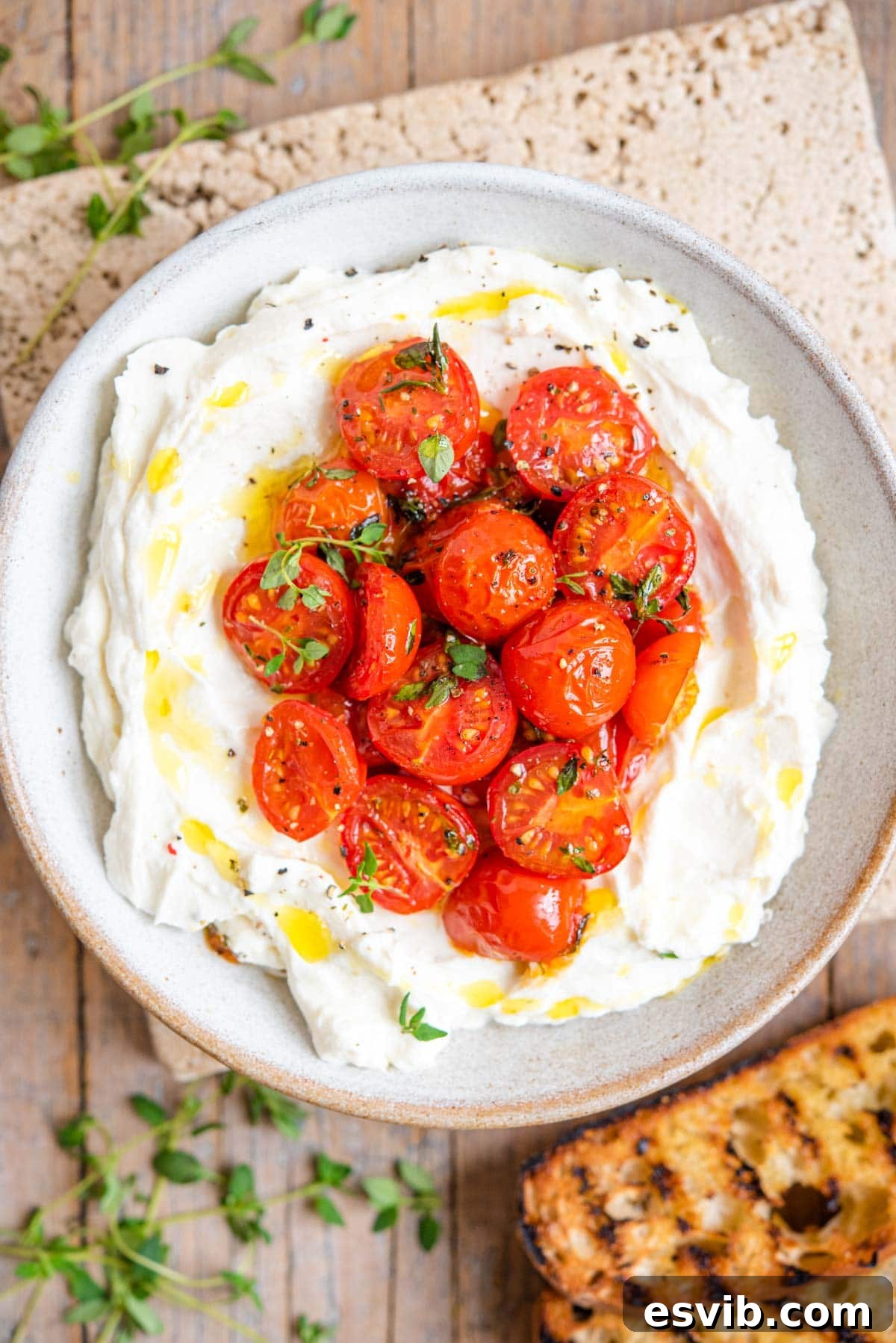 An overhead shot of ricotta in a serving bowl topped with roasted tomatoes, garnished with fresh herbs and a drizzle of olive oil.